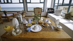 Una mesa de desayuno con diferentes platos, un florero con una sola flor y vista al océano a través de amplios ventanales.