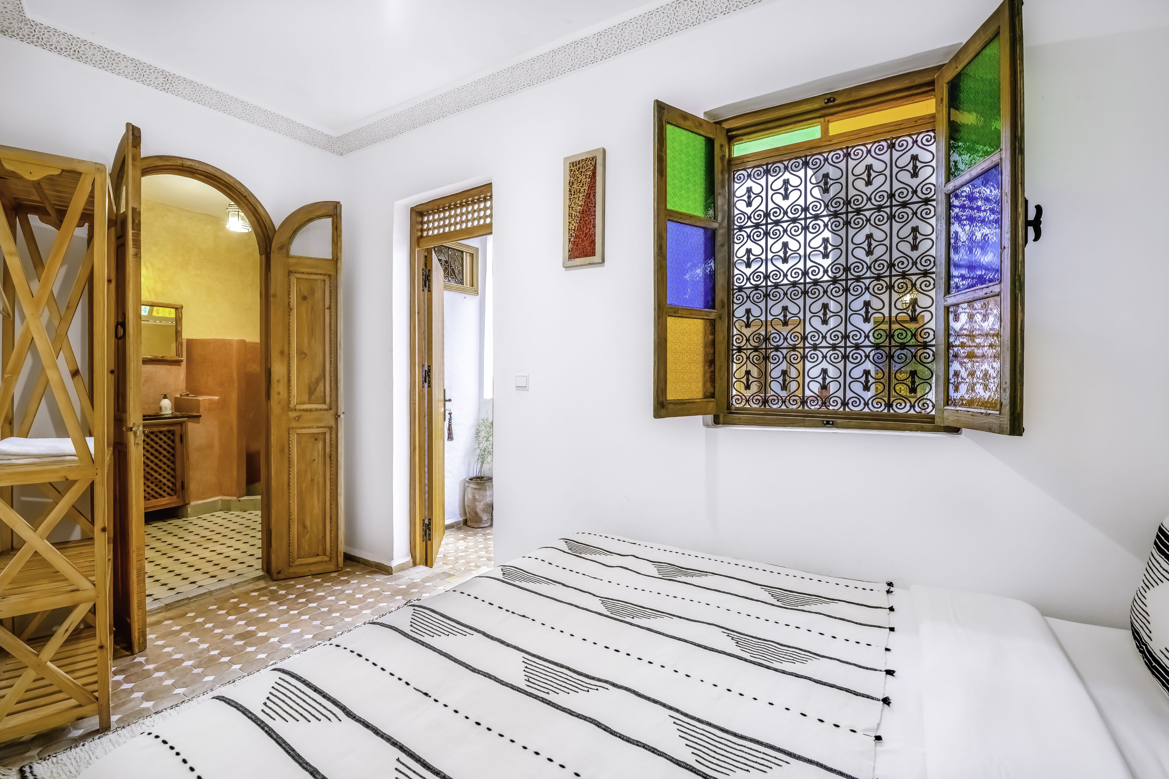 A room with a bed, a wooden door, a window with stained glass, and a patterned rug.