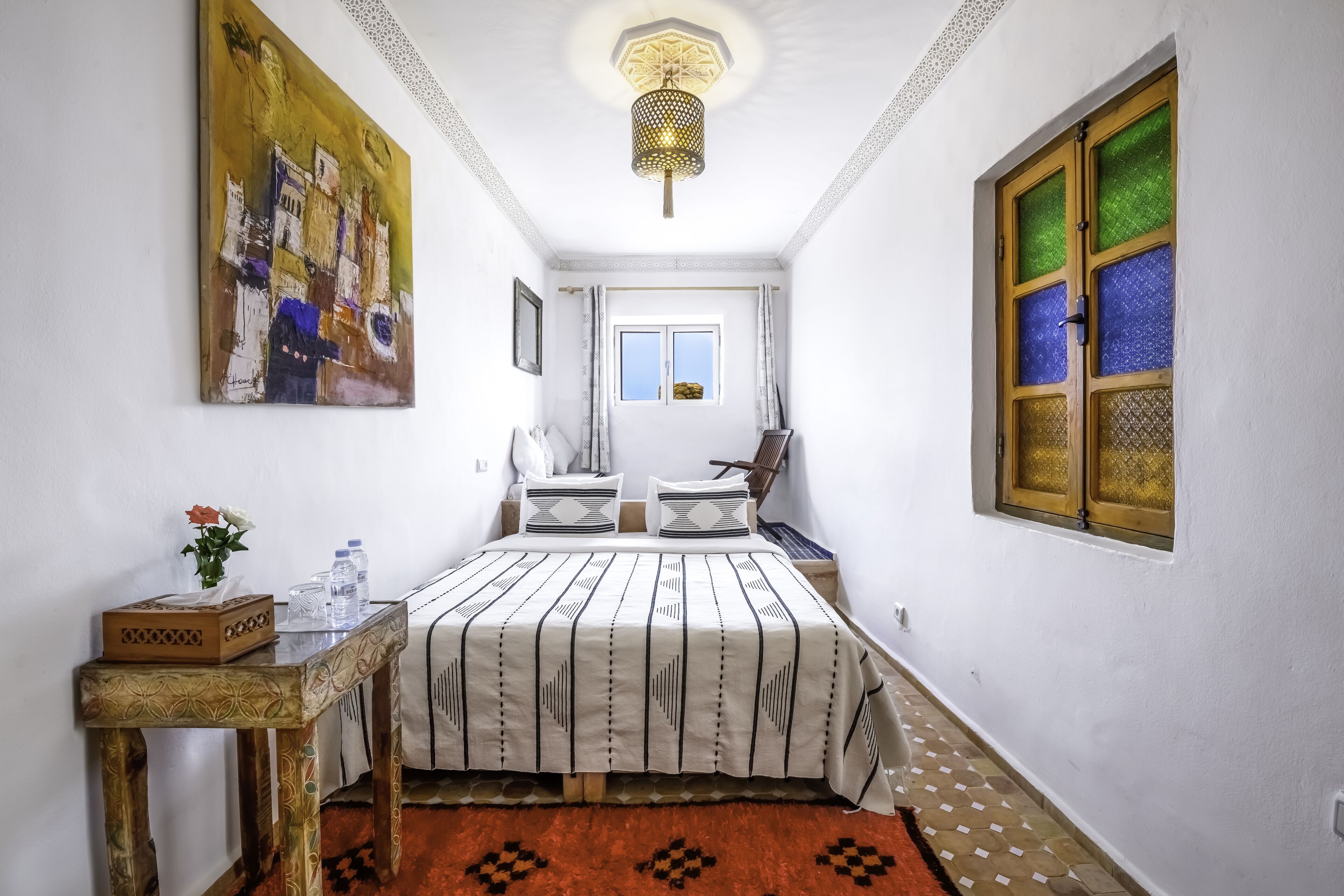 Un dormitorio con cama, una mesita de noche, una mesa con un jarrón y botellas, un cuadro en la pared y una ventana con vidrio ornamental.