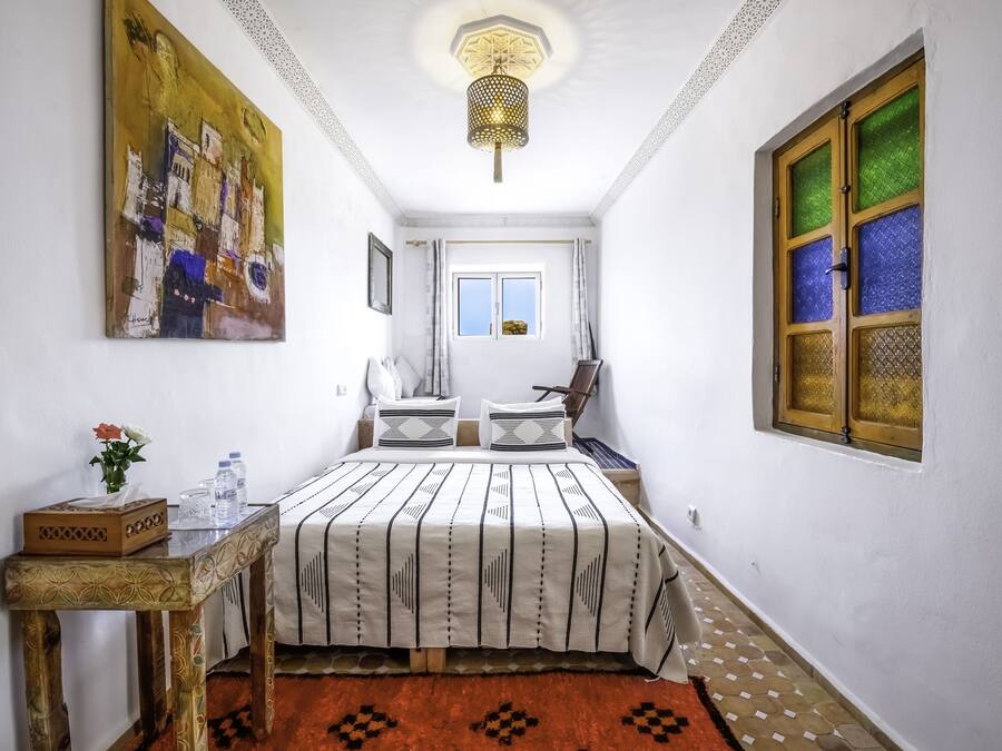 Un dormitorio con cama, una mesita de noche, una mesa con un jarrón y botellas, un cuadro en la pared y una ventana con vidrio ornamental.