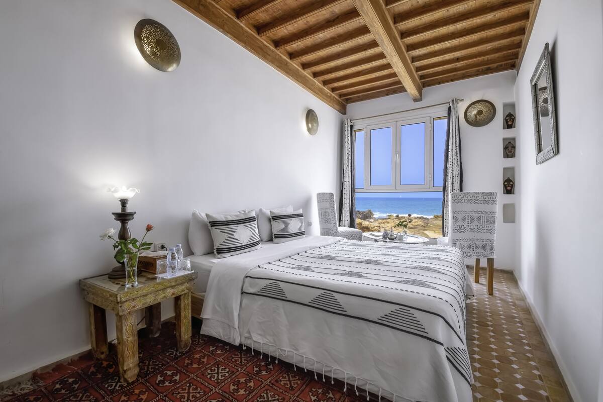Un dormitorio con una cama grande, una mesita de noche con lámpara y vistas al mar a través de una ventana.