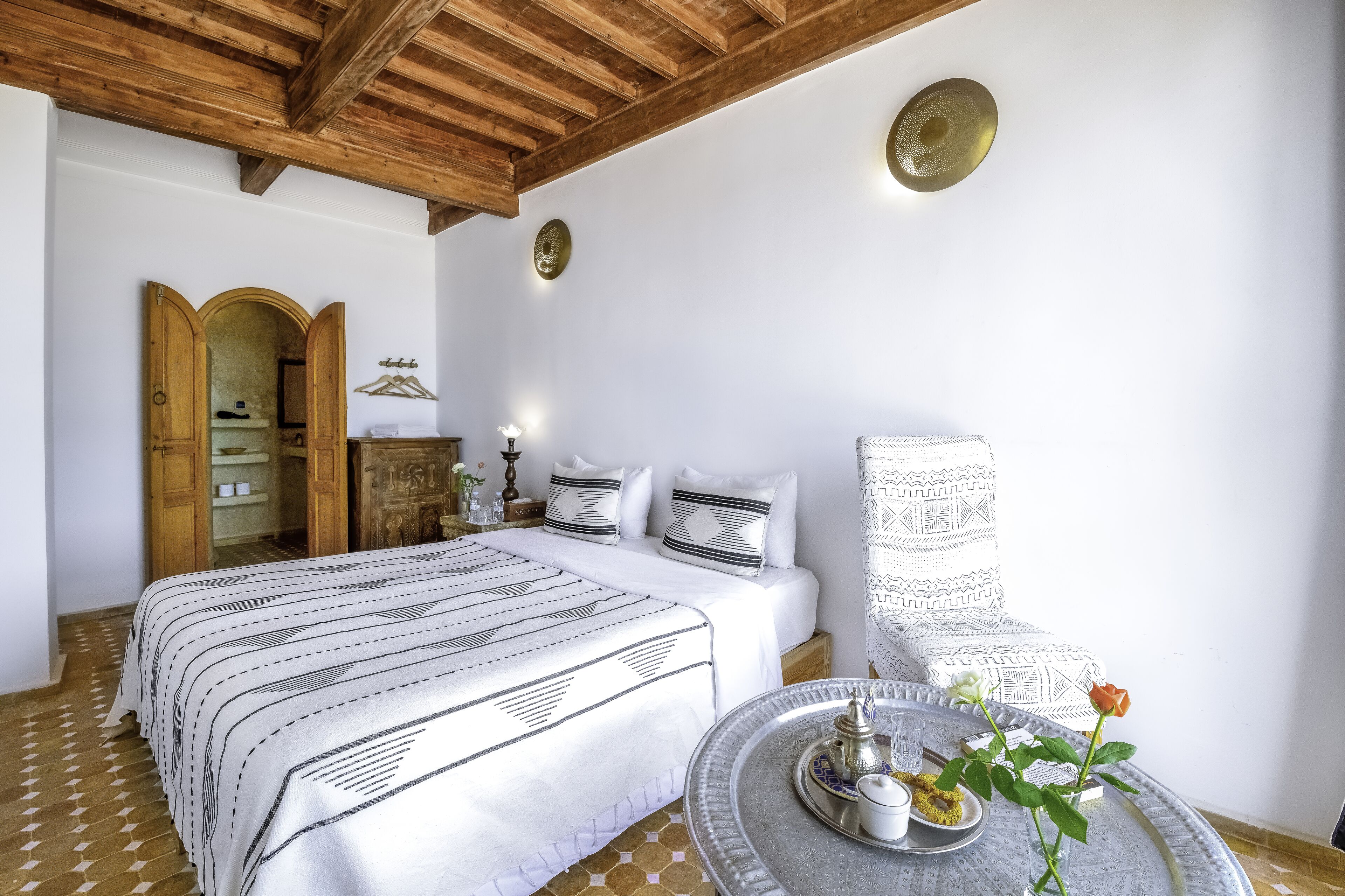 Un dormitorio con una cama grande, vigas de techo de madera, una mesita redonda con una bandeja y un estante montado en la pared.