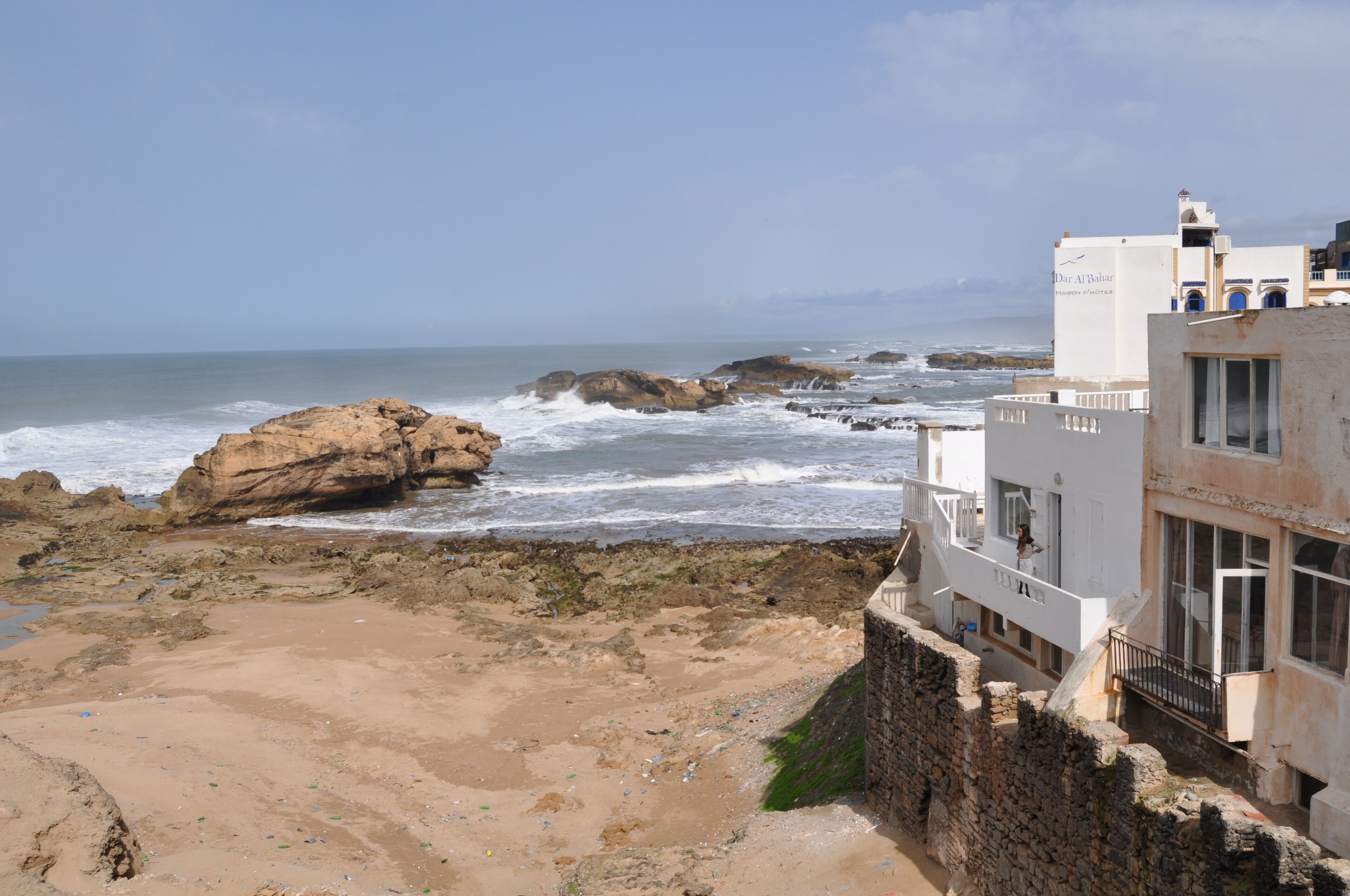 A coastal building with a balcony overlooking the sea, a rocky shoreline, and a clear sky.