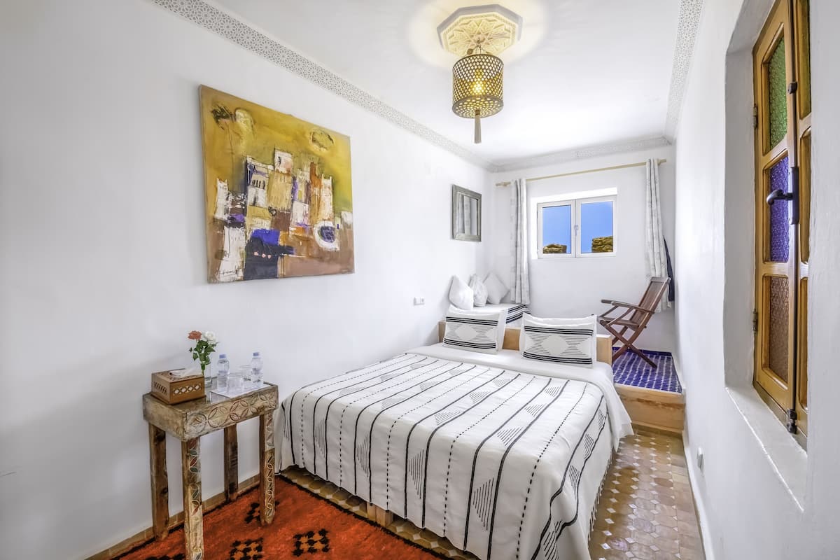 Un dormitorio con cama, mesita de noche, una silla, ventana y un cuadro en la pared.