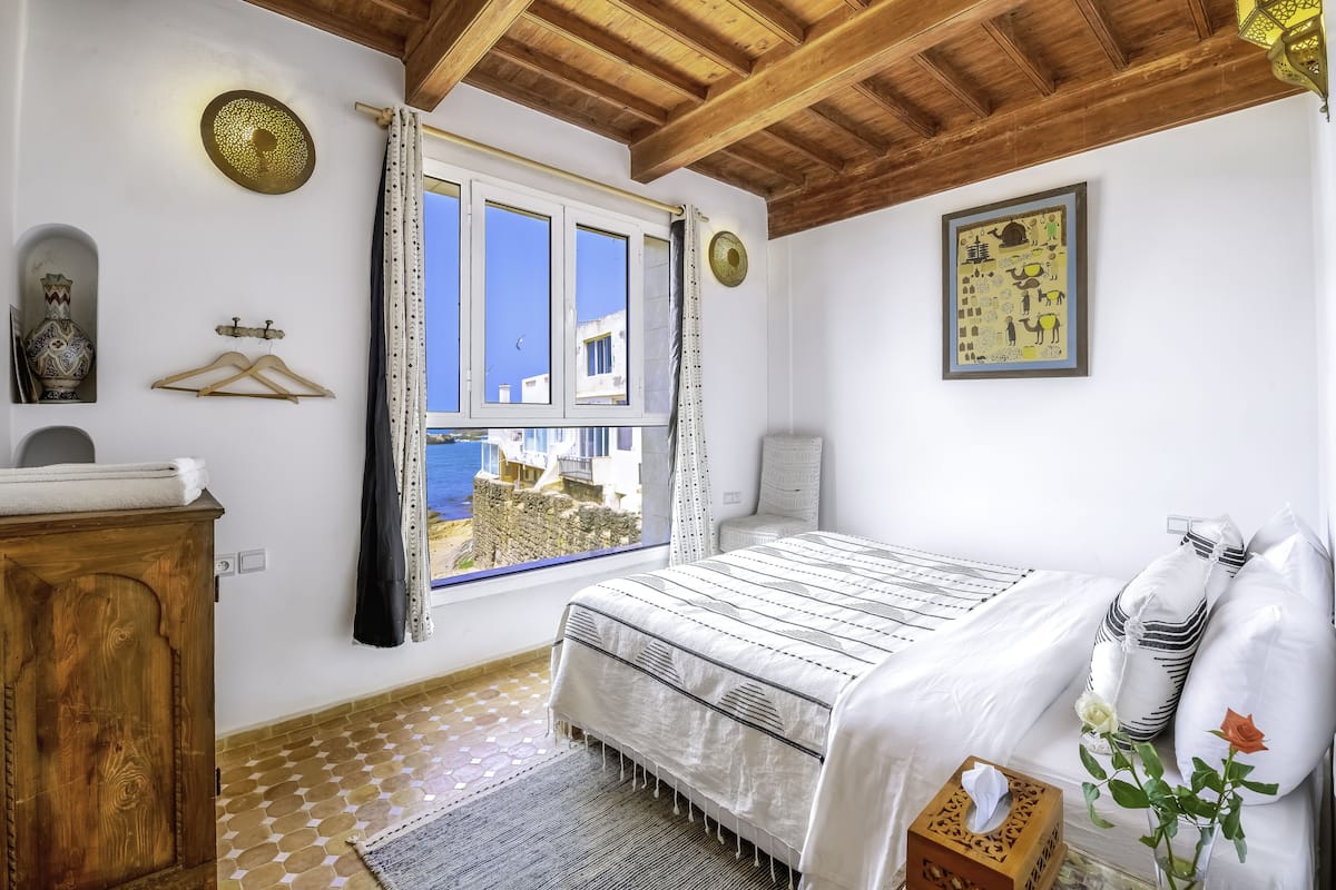 Un dormitorio con cama, techo de madera, ventana con vistas, cómoda de madera y mesita de noche con caja de pañuelos y flores.