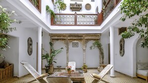 Un patio con techo alto, paredes blancas y un balcón de madera. Hay plantas en macetas, una mesa con sillas y un sofá.