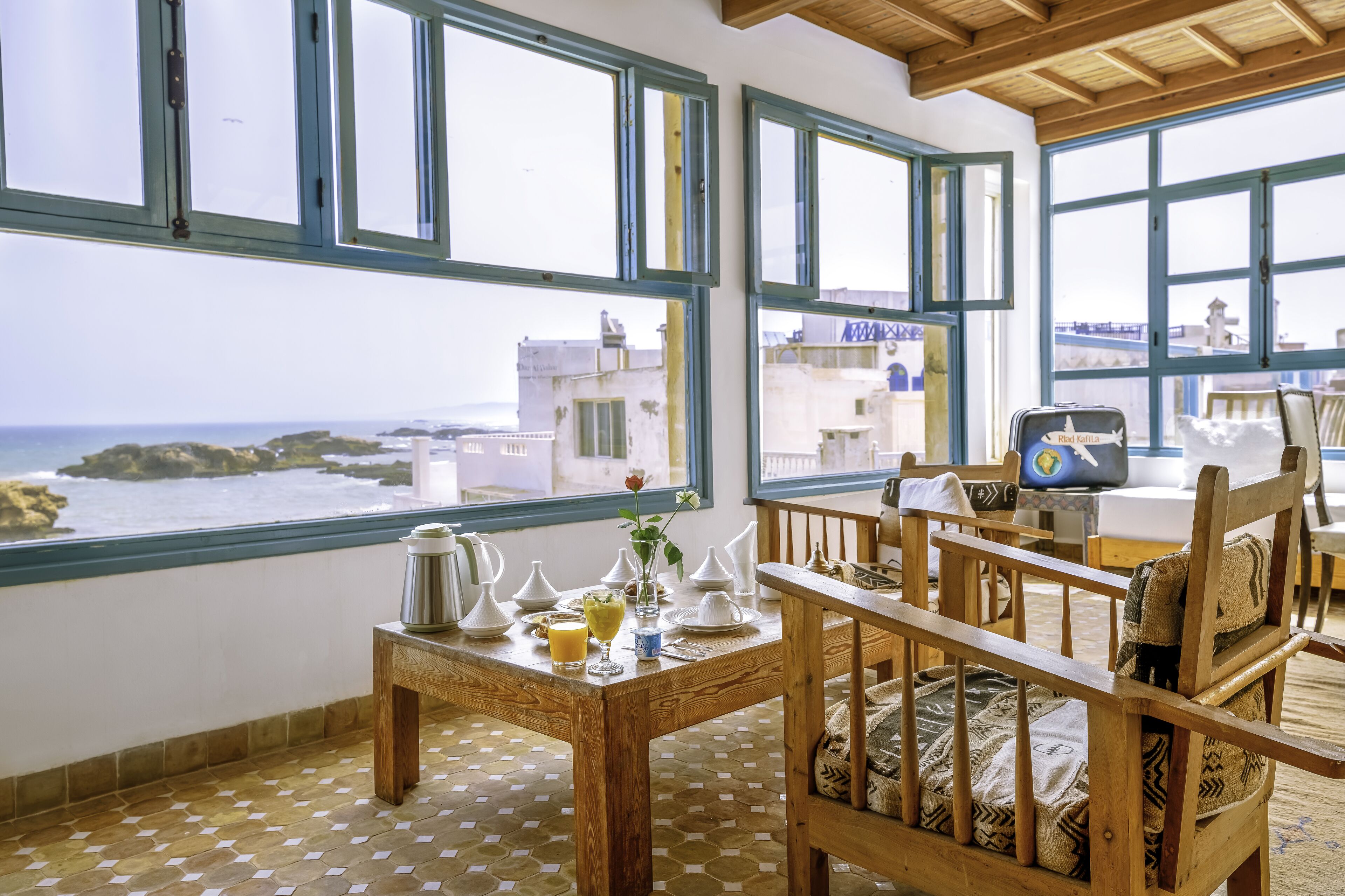 A room with large windows overlooking the sea, a wooden table with a coffee pot, and a chair with a patterned cushion.