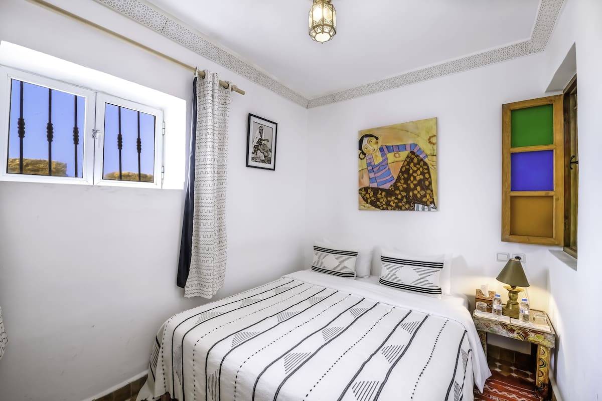 Un dormitorio con cama, mesita de noche, lámpara, ventana con cortinas y un cuadro en la pared.
