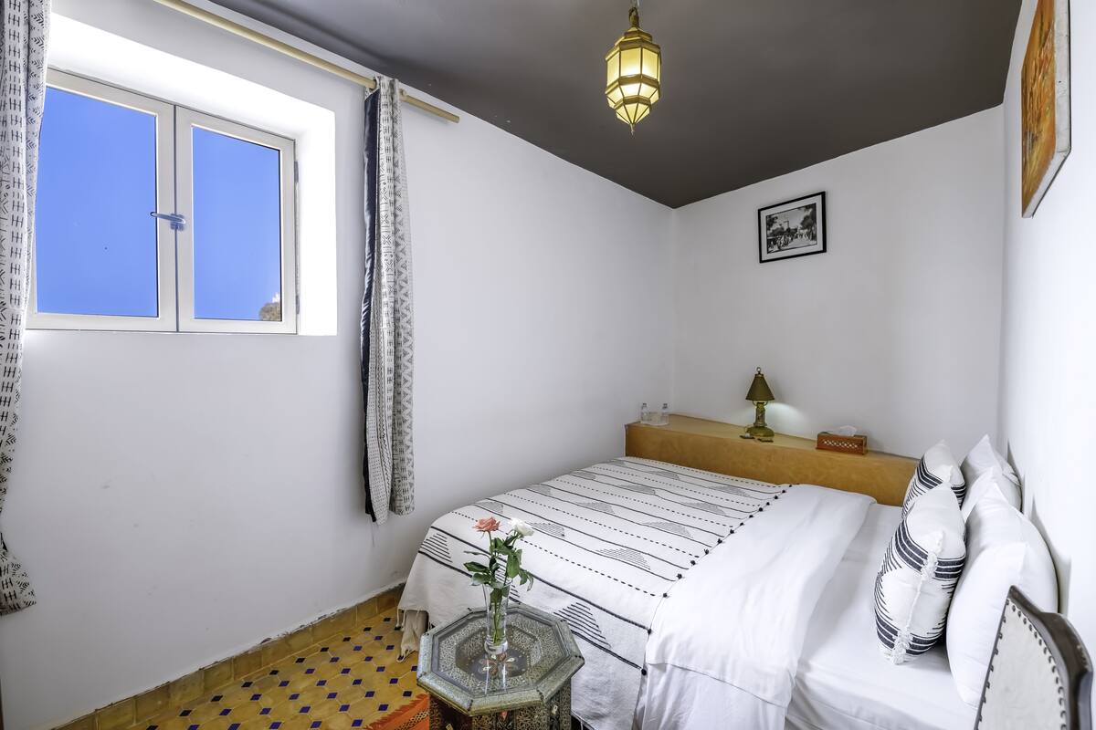Un dormitorio con una cama, una mesita de noche, una ventana con cortinas, una lámpara colgante y un cuadro en la pared.
