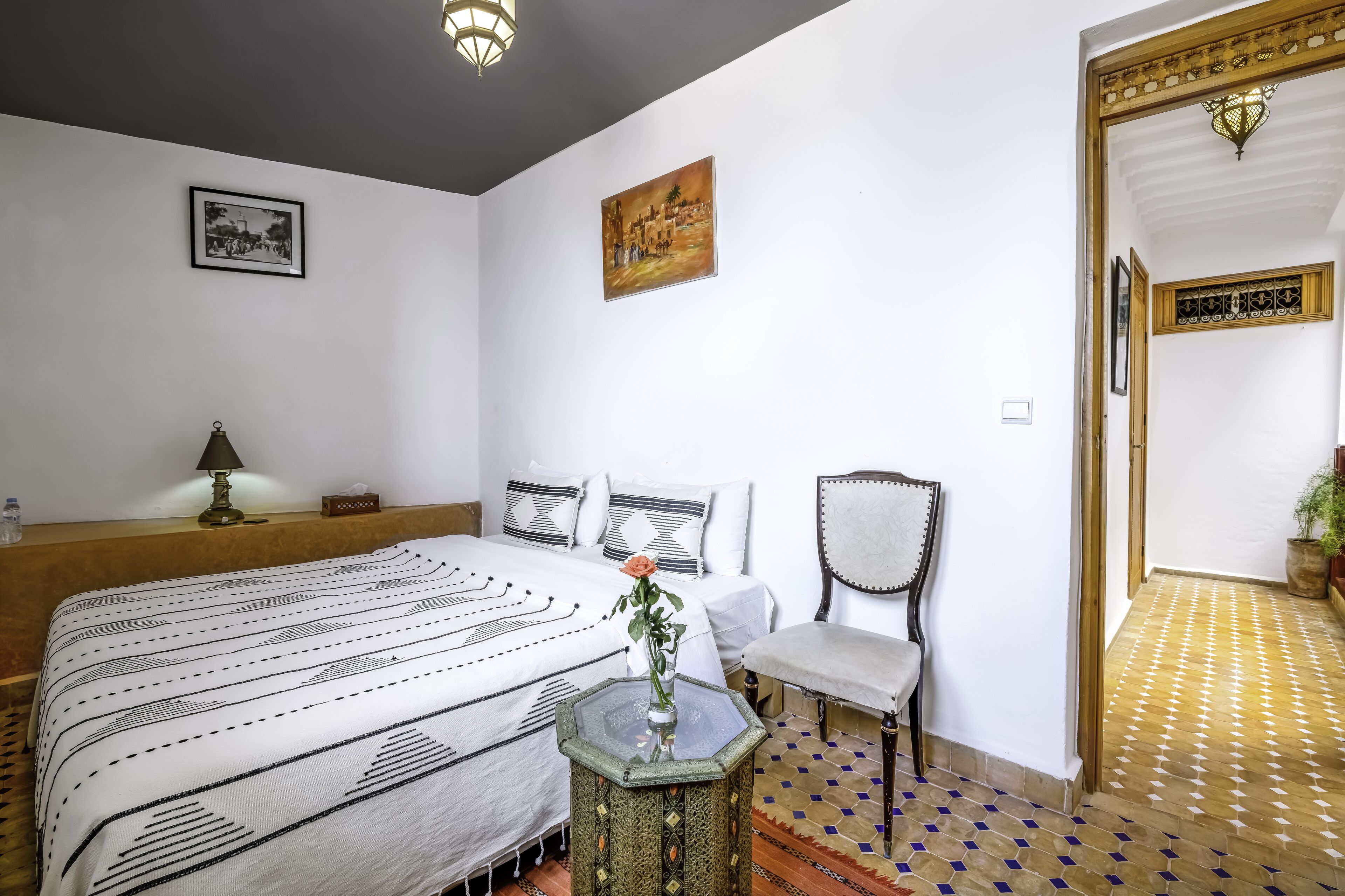 A bedroom with a large bed, a chair, a small table with a vase, and a painting on the wall.