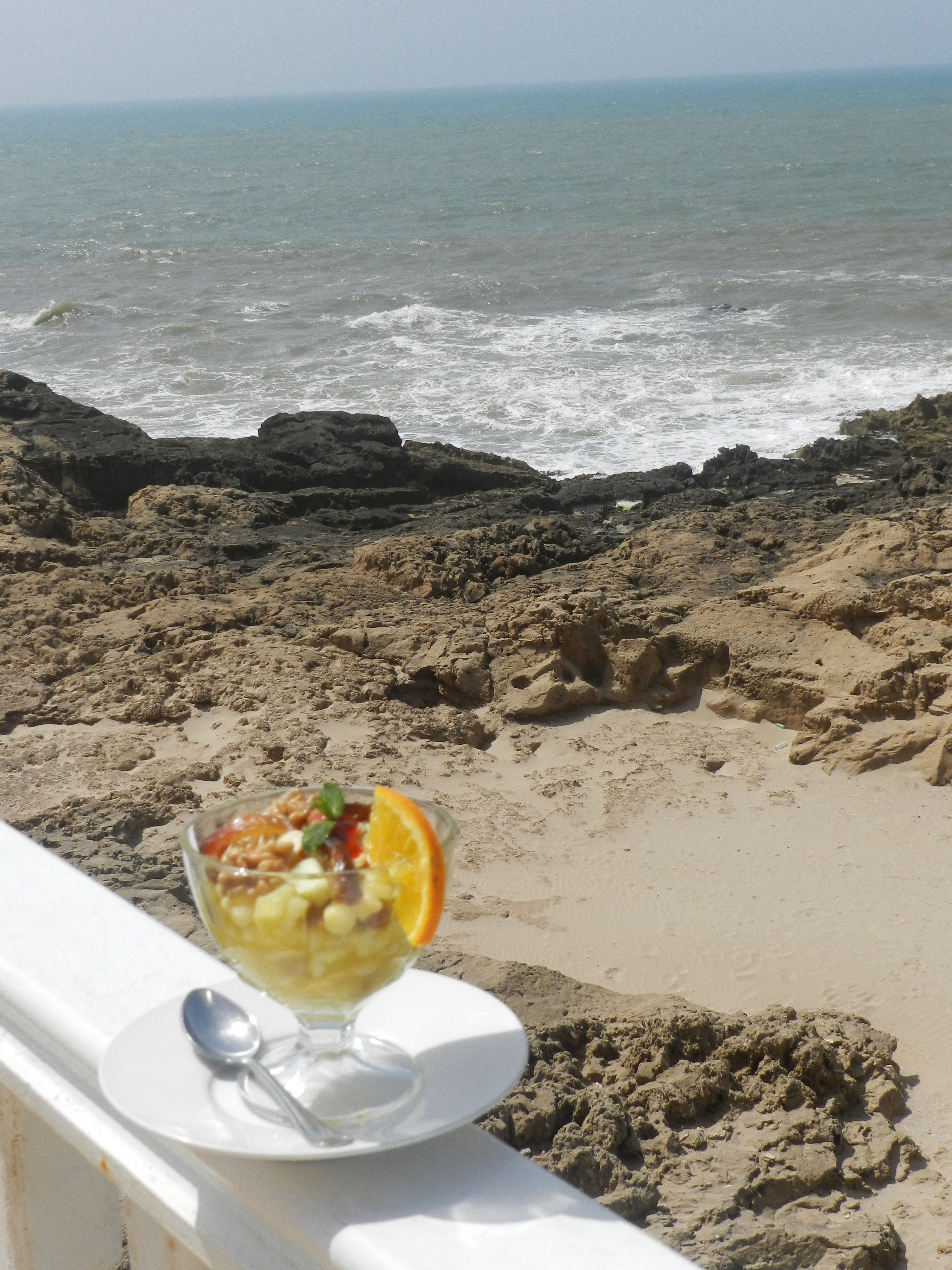 Un postre en un plato blanco, con una cuchara, con vista a una playa rocosa y al océano.