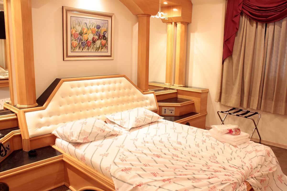 A hotel room with a large bed, a bedside table, a nightstand, a mirror, a painting on the wall, and a window with curtains.