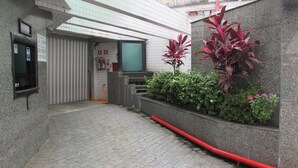 Pasillo de hotel con suelo de baldosas, mostrador de recepción, extintor y una maceta con follaje rojo.
