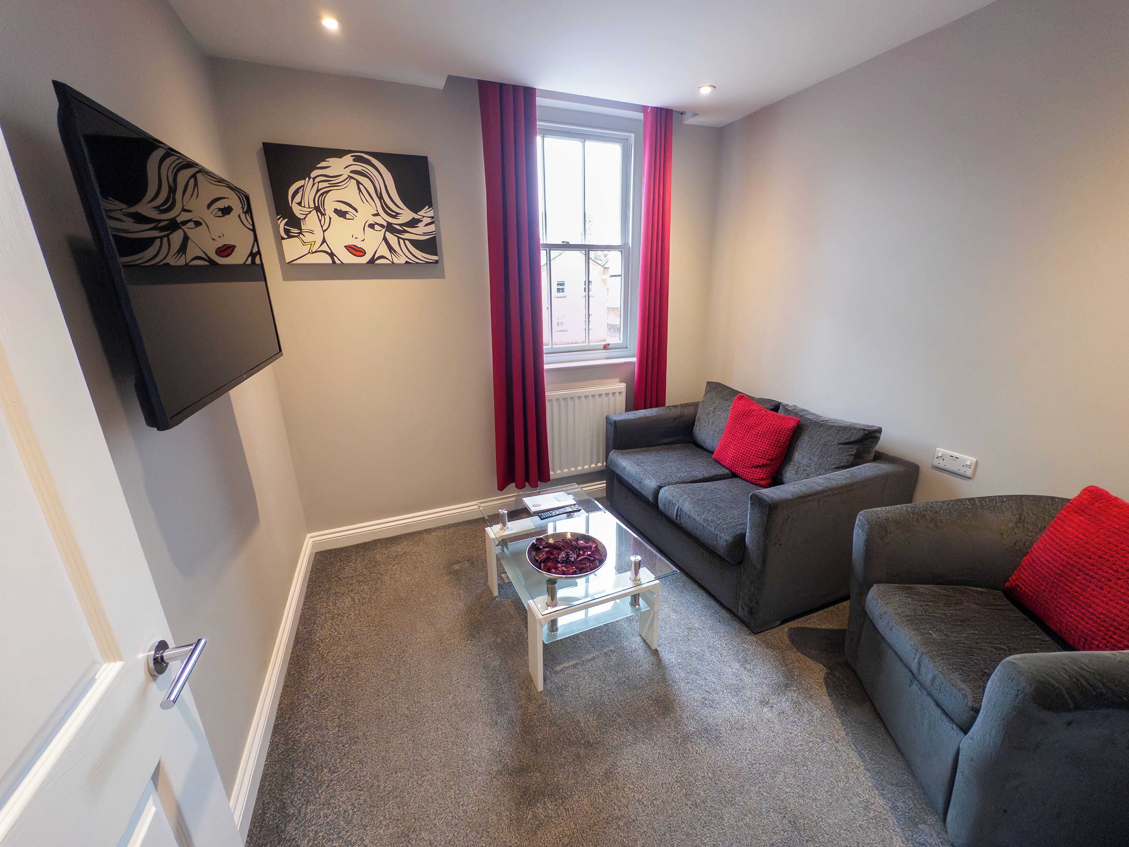 A modern living room with a grey sofa, a glass coffee table, and red cushions.