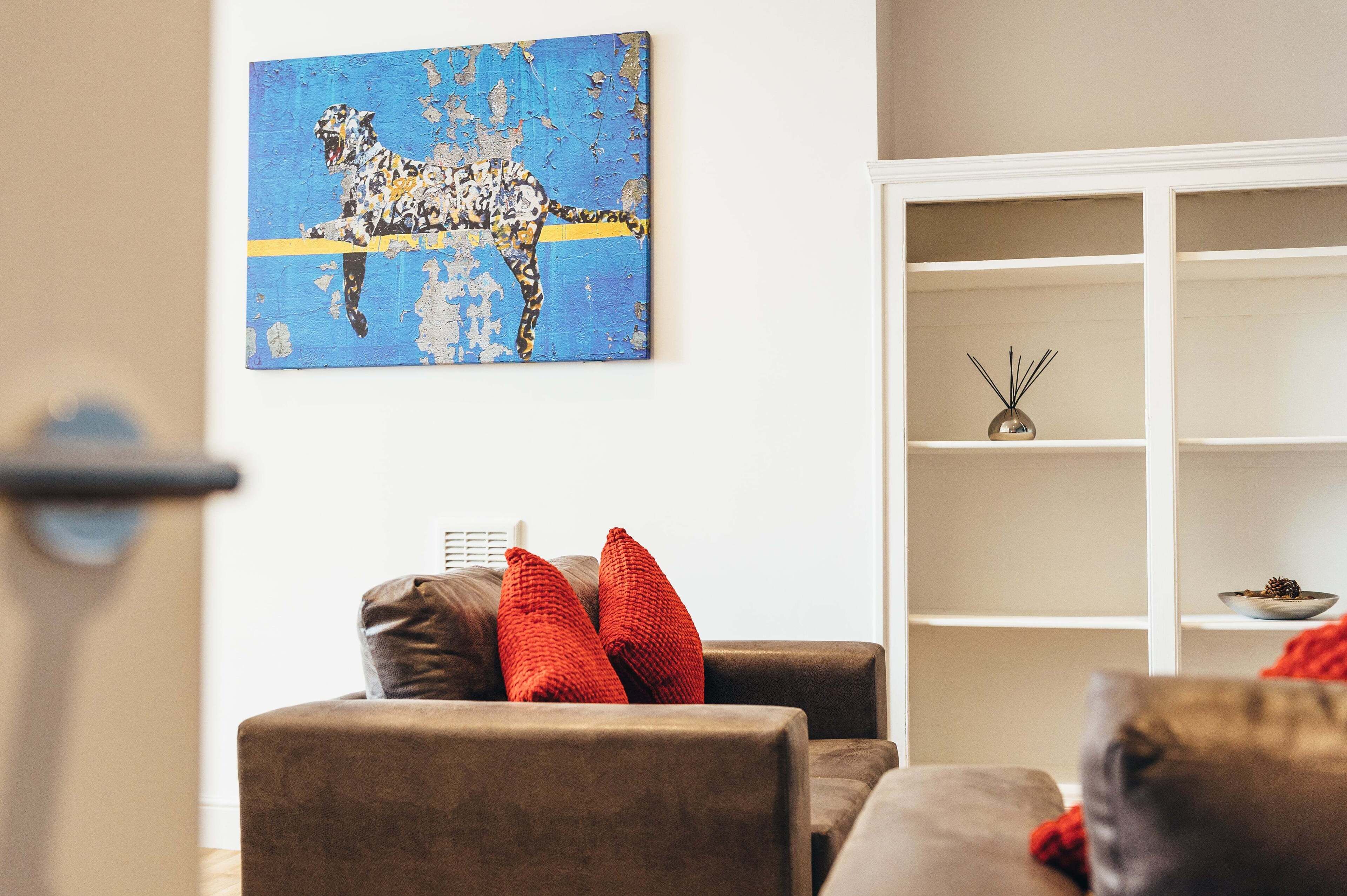 A living room with a sofa, red cushions, a wall-mounted painting of a giraffe, and a white shelving unit with decorative items.