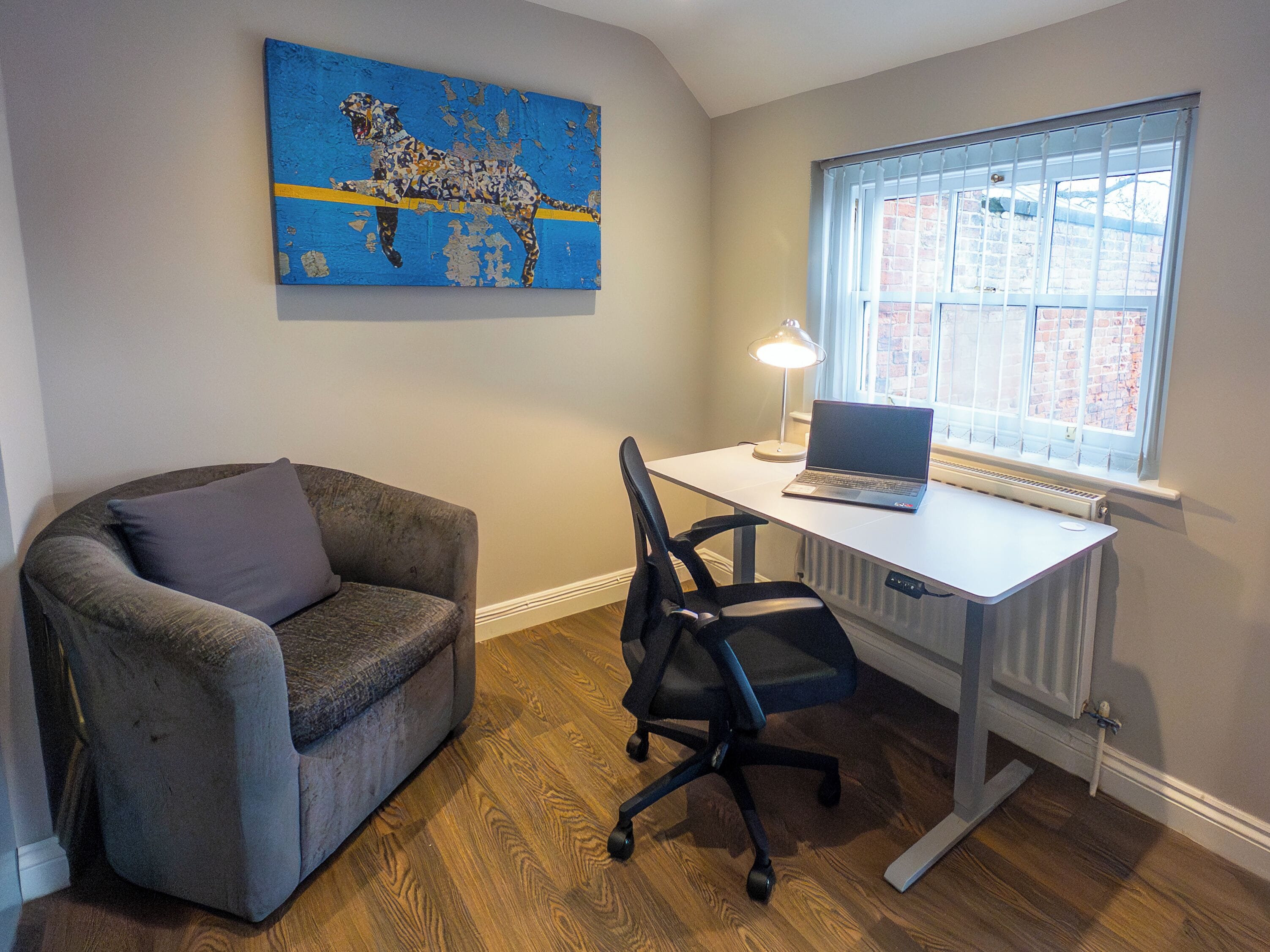 A room with a grey armchair, a black office chair, a white desk with a laptop, a lamp, and a radiator.