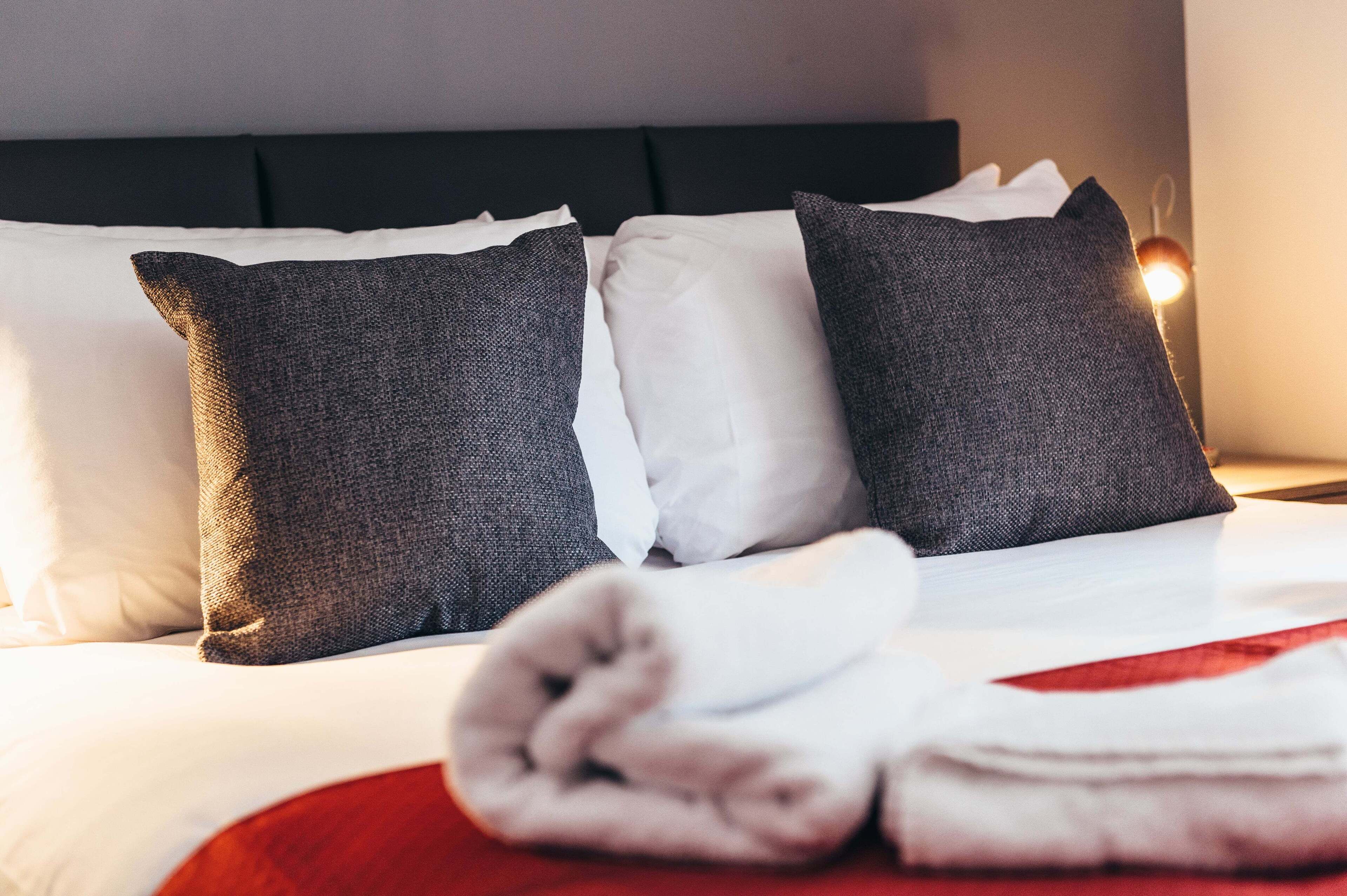 A neatly made bed with white and grey pillows, a folded towel, and a red blanket.