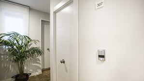 A hallway with a potted plant, a door with a number plate, and a wall-mounted intercom. - Hola Rooms (Madrid)