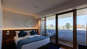 Une chambre d’hôtel moderne dotée d’un grand lit, d’une table de chevet, d’un luminaire fixé au mur et d’une porte-fenêtre coulissante donnant sur un balcon extérieur.
