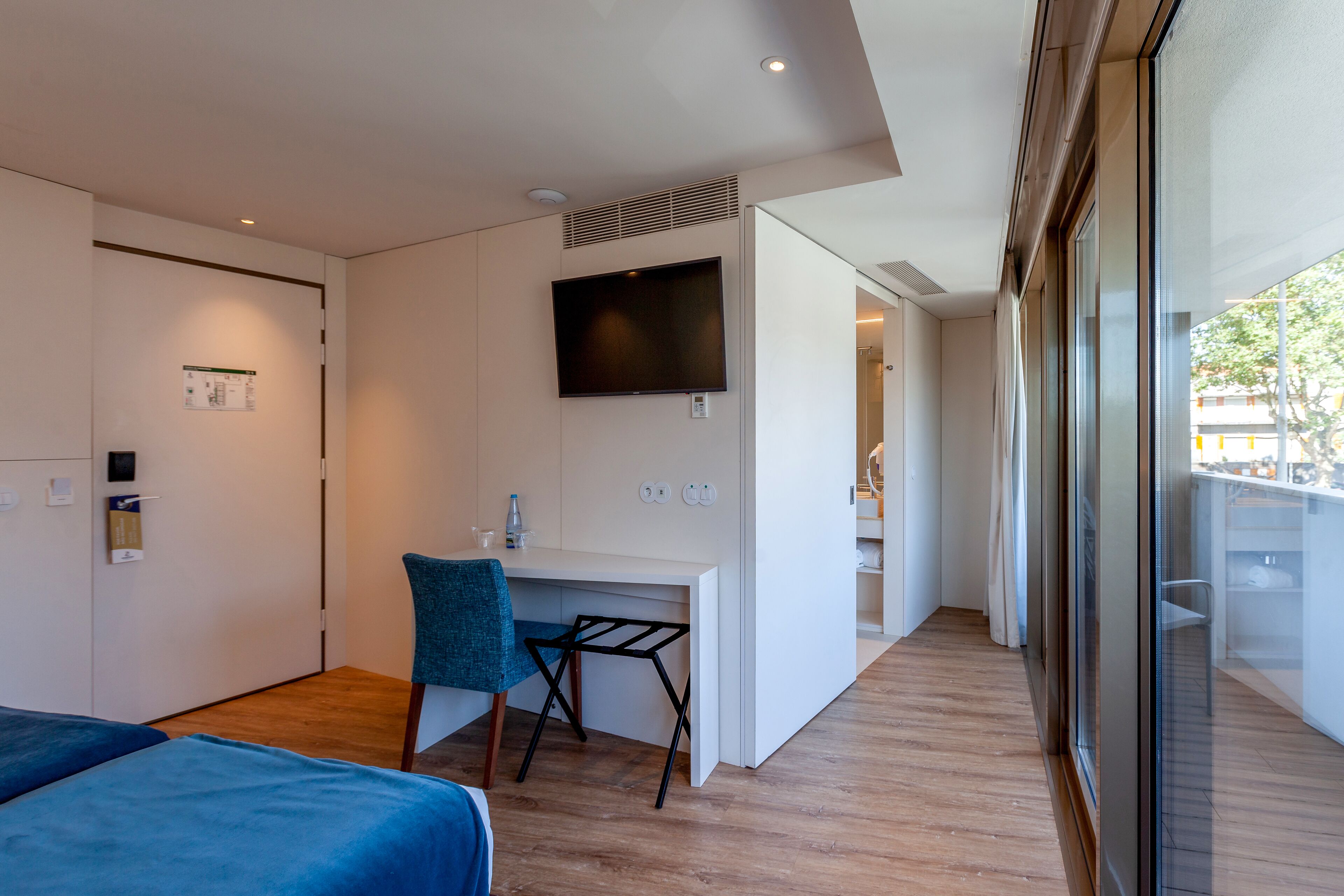A modern hotel room with a bed, a desk, a chair, a TV, and a large window.