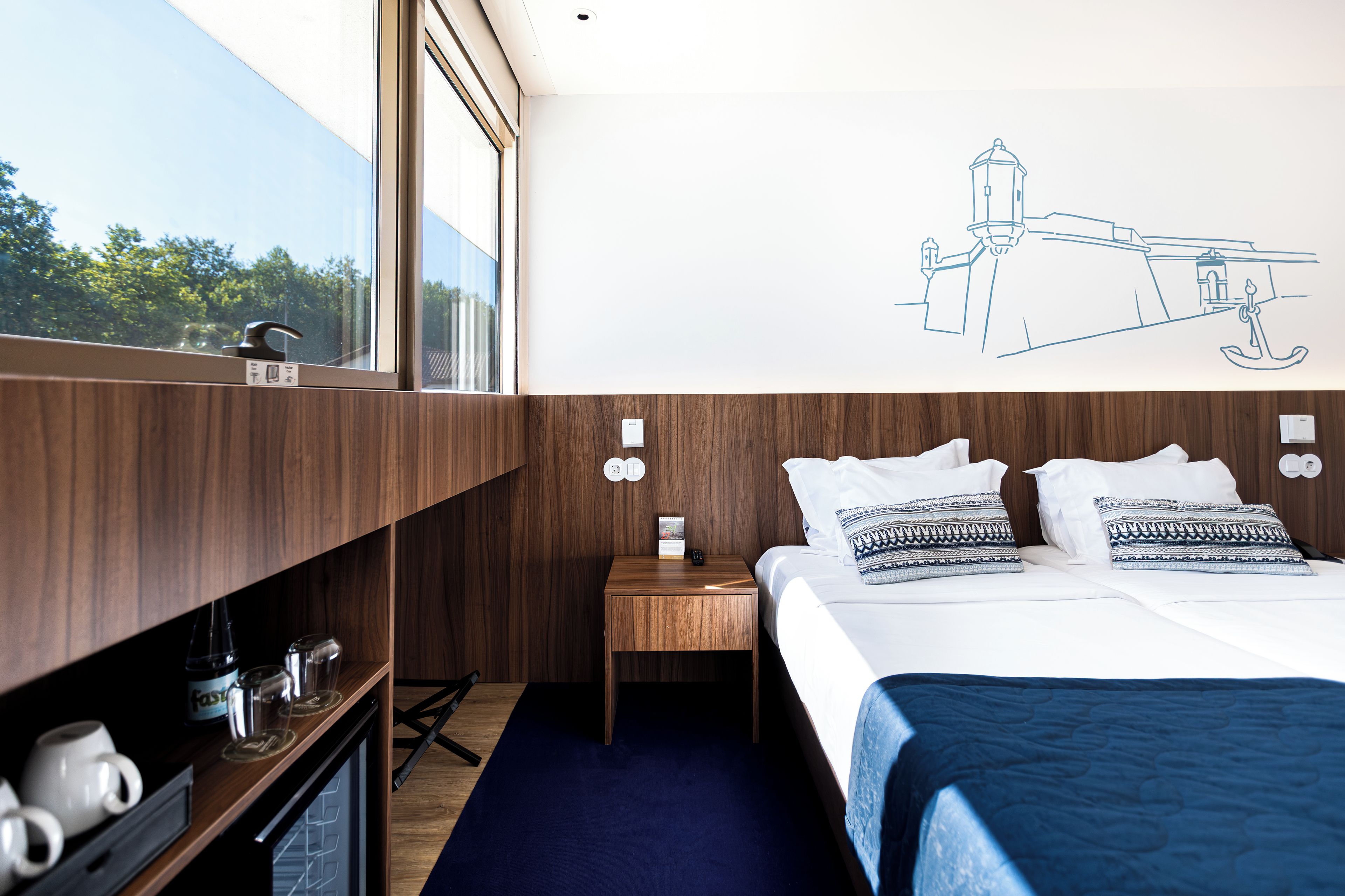 A hotel room with a large window, a bed with patterned pillows, a nightstand, and a wall-mounted light fixture.