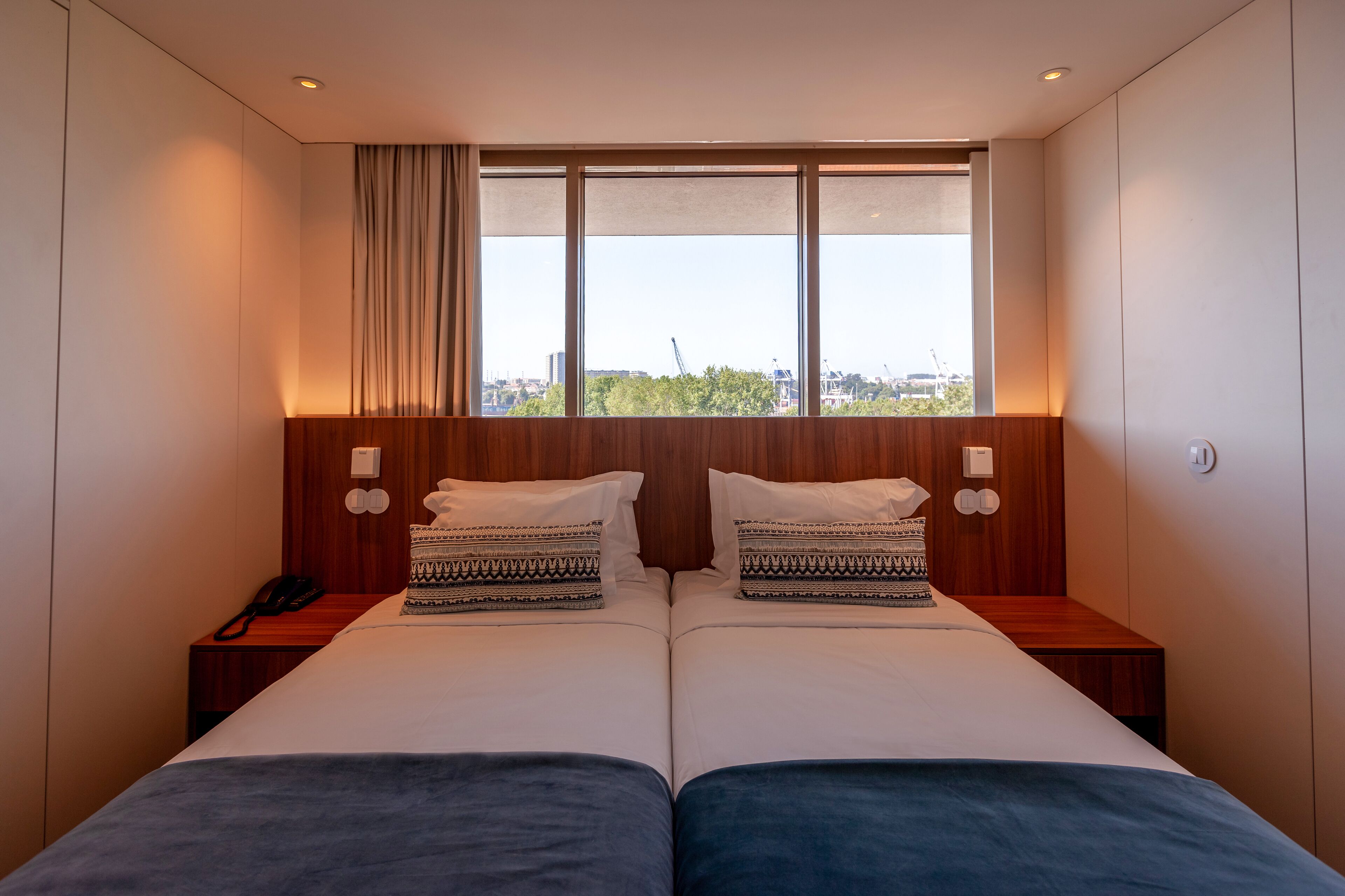A double bed with patterned pillows, a bedside phone, and a window with a view of buildings and trees.