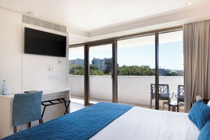 Habitación de hotel con cama, televisor montado en la pared, escritorio con silla, una pequeña mesa con otra silla, y un balcón con vistas a la ciudad.