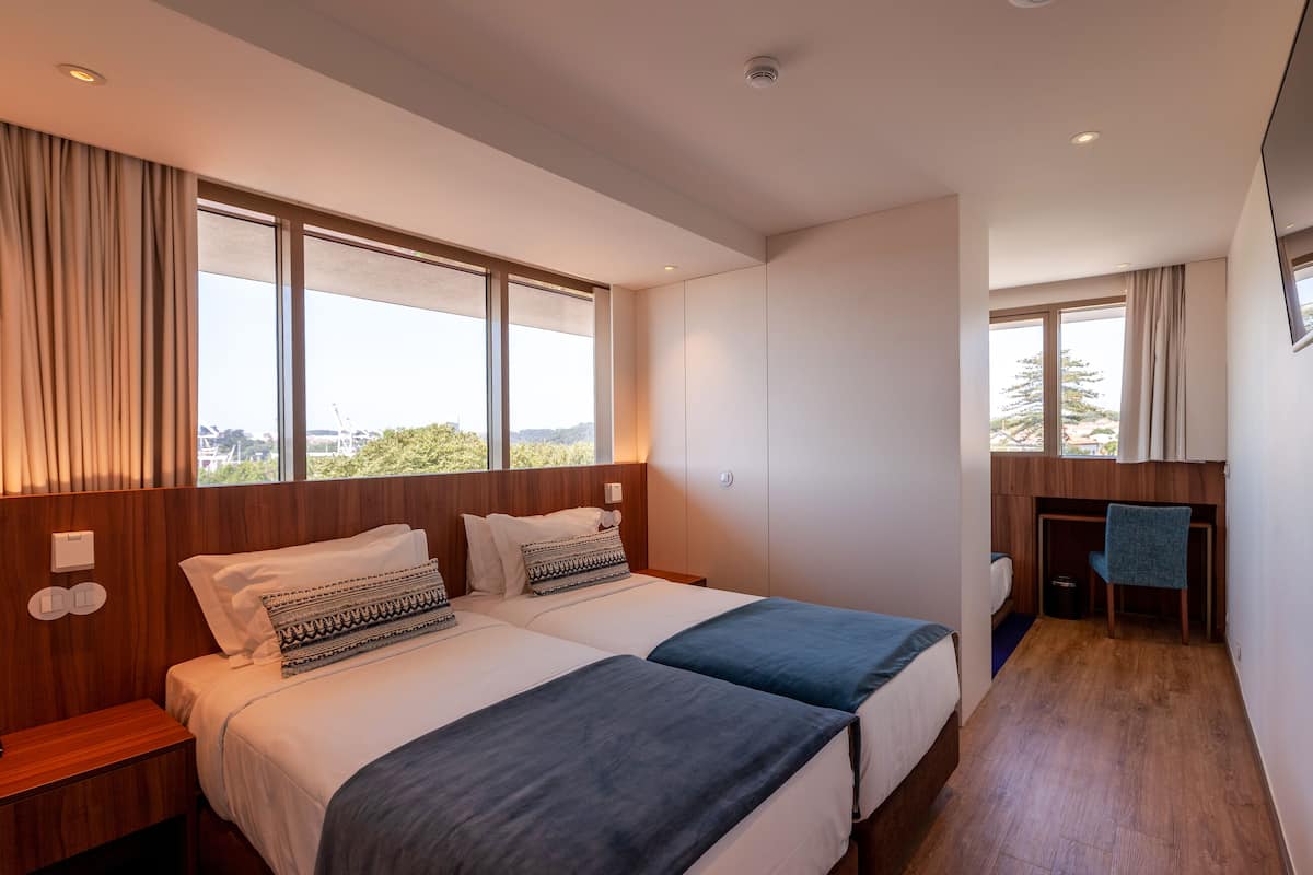A modern hotel room with two beds, a desk, and large windows offering a city view.