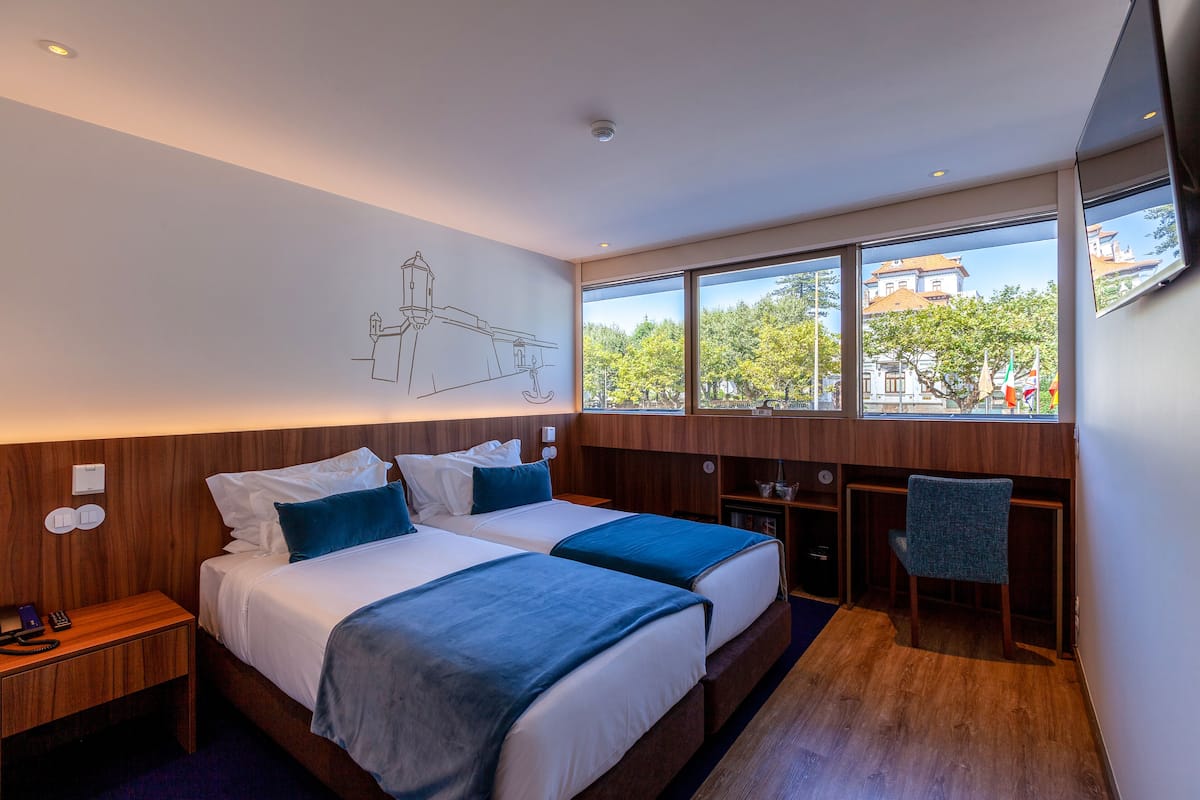 A hotel room with two beds, a desk, and a chair. There is a large window with a view of buildings and trees.