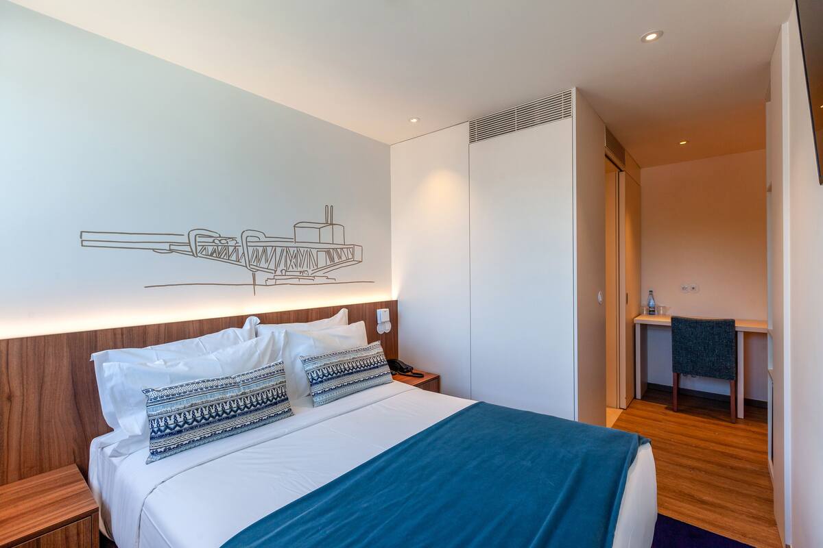 A modern hotel room with a large bed, a desk, and a wardrobe.