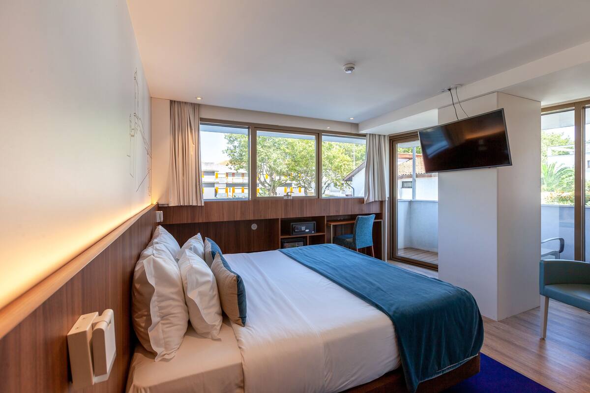 A modern hotel room with a large bed, a flat-screen TV, a desk, and a view of the outdoors.