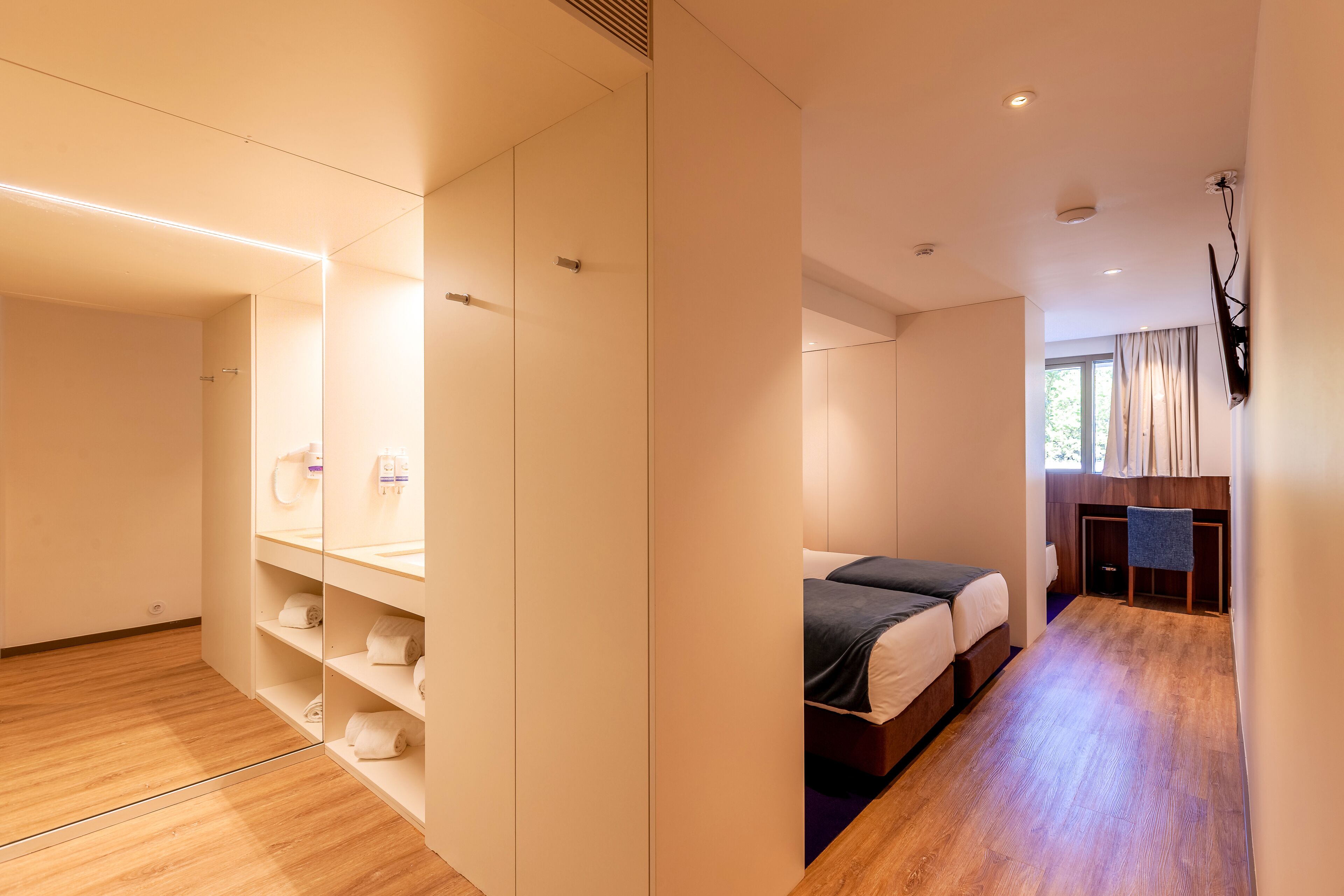 Una habitación de hotel moderna con piso de madera, un armario espacioso, una cama con una manta oscura y un escritorio con una silla.