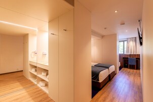 Una habitación de hotel moderna con piso de madera, un armario espacioso, una cama con una manta oscura y un escritorio con una silla.