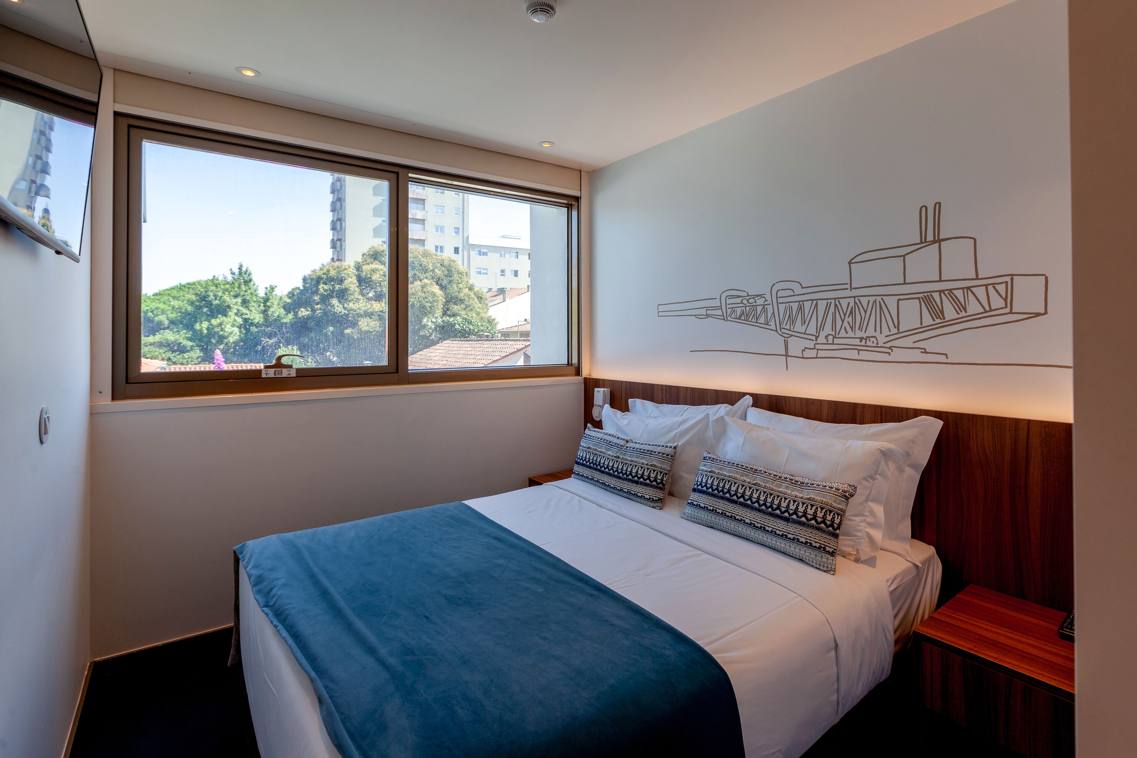A hotel room with a large bed, a window with a view of buildings, and a wall with a simple line drawing.