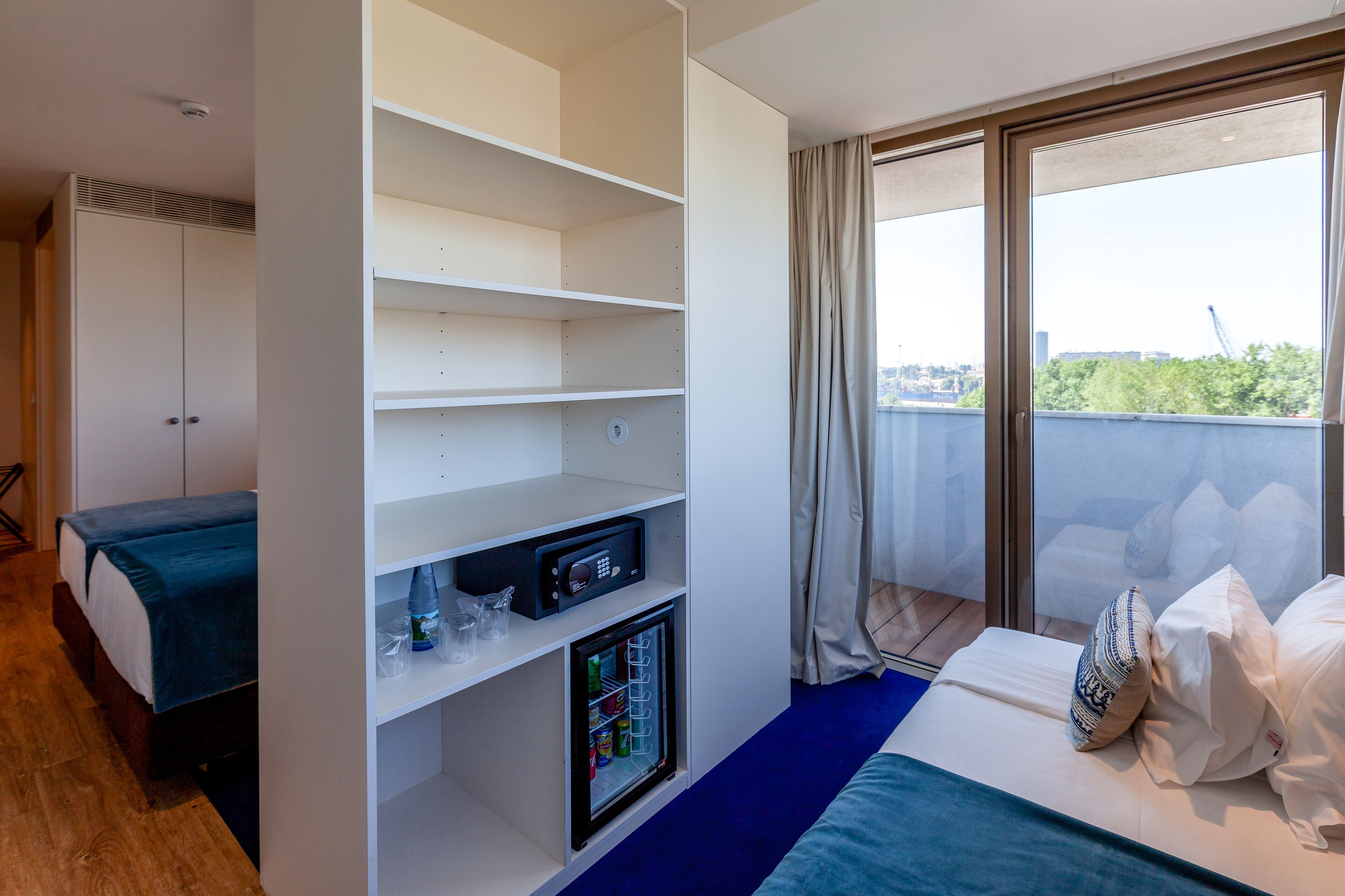 A hotel room with a bed, a mini-fridge, a shelf unit, and a view of the city through large windows.