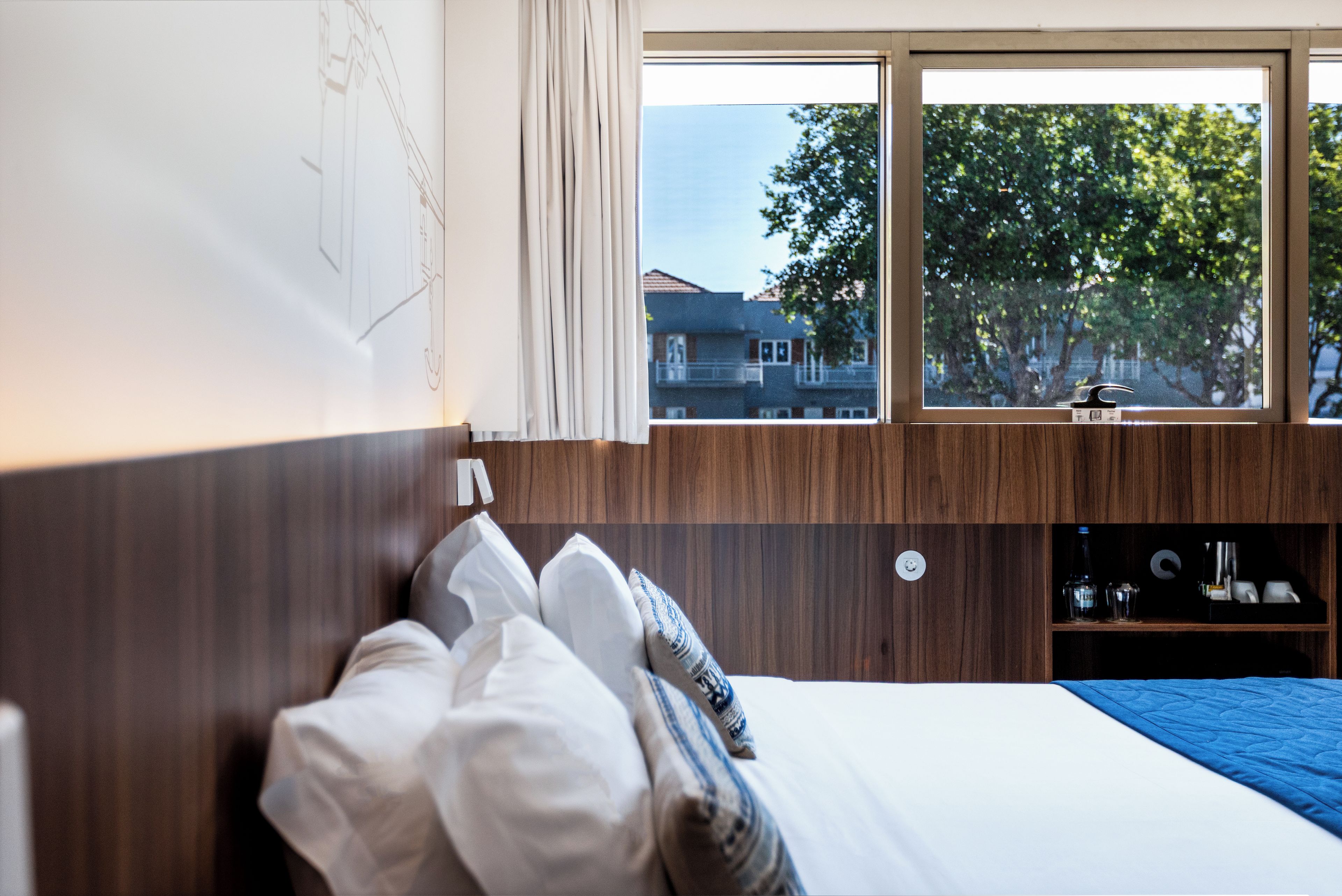 A hotel room with a bed, a window with a view of trees and buildings, and a wooden headboard.