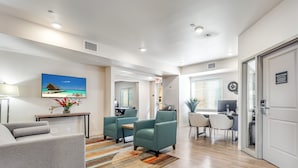 Un salon spacieux avec un canapé gris, des fauteuils turquoise et un tapis coloré. On y trouve également un coin repas avec une table et des chaises, et une cuisine à l’arrière-plan.