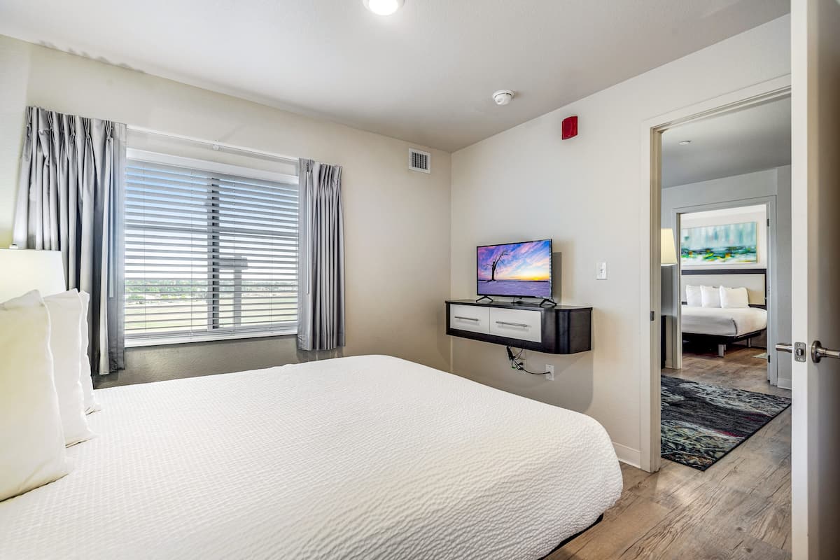 A hotel room with a large bed, a flat-screen TV mounted on the wall, a bedside table, and a window with blinds.