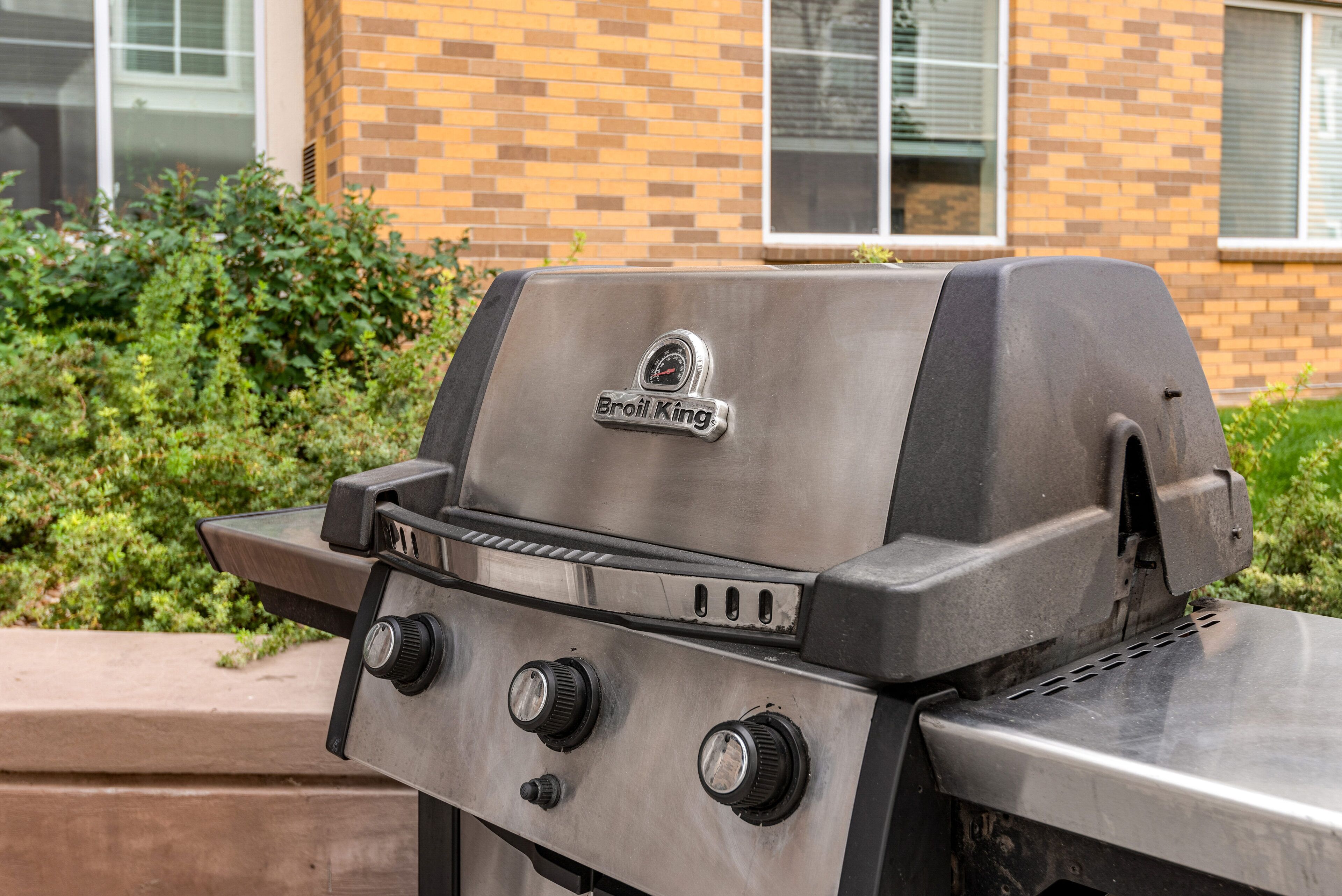 A stainless steel grill with a digital thermometer and control knobs, set against a brick building and greenery.
