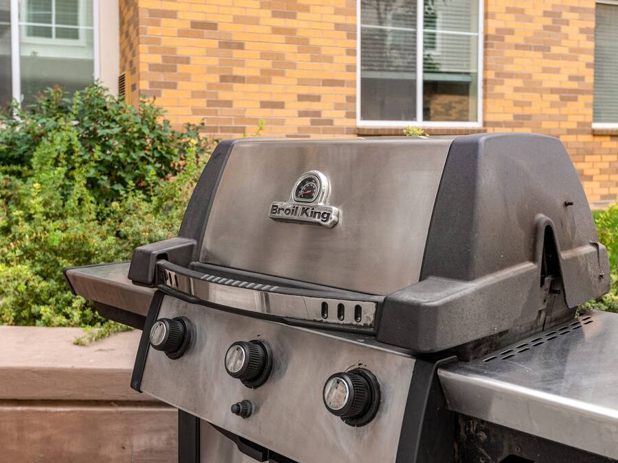 A stainless steel grill with a digital thermometer and control knobs, set against a brick building and greenery.