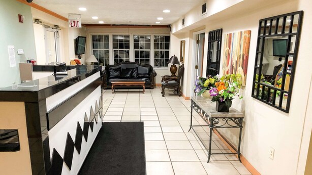 A hotel lobby with a reception desk, a black leather sofa, a coffee table, a small table with flowers, and a large window with blinds. - Knights Inn Lake City (Lake City)