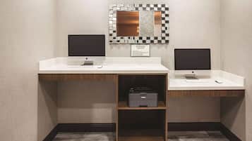Un centre d’affaires équipé de deux ordinateurs Apple iMac sur des bureaux blancs, d’une imprimante Brother sur une étagère et d’un miroir avec un cadre à carreaux.