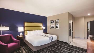 A hotel room with a large bed, a nightstand, a chair, a desk, and a door leading to another room.