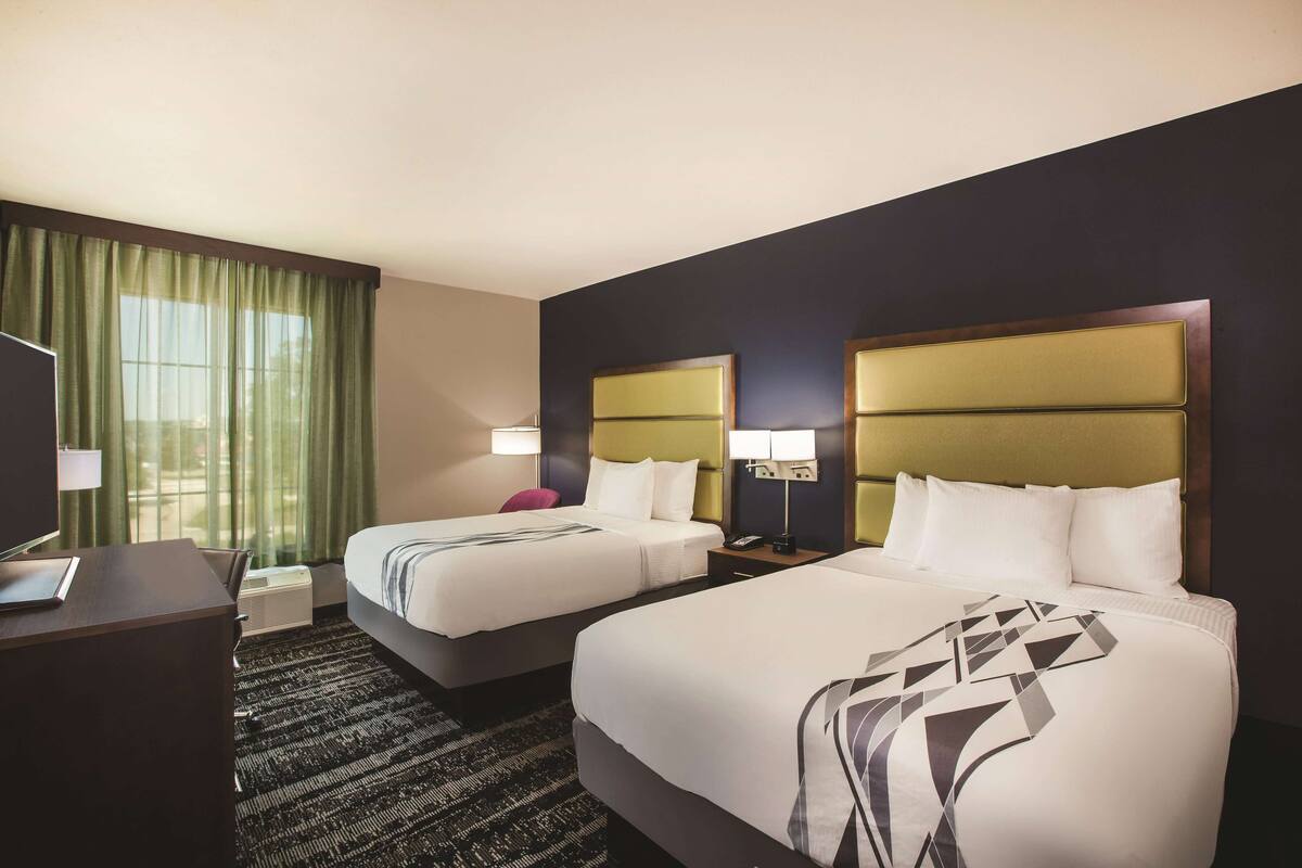 Une chambre d’hôtel avec deux lits, un bureau, une chaise, une lampe et une fenêtre avec des rideaux.