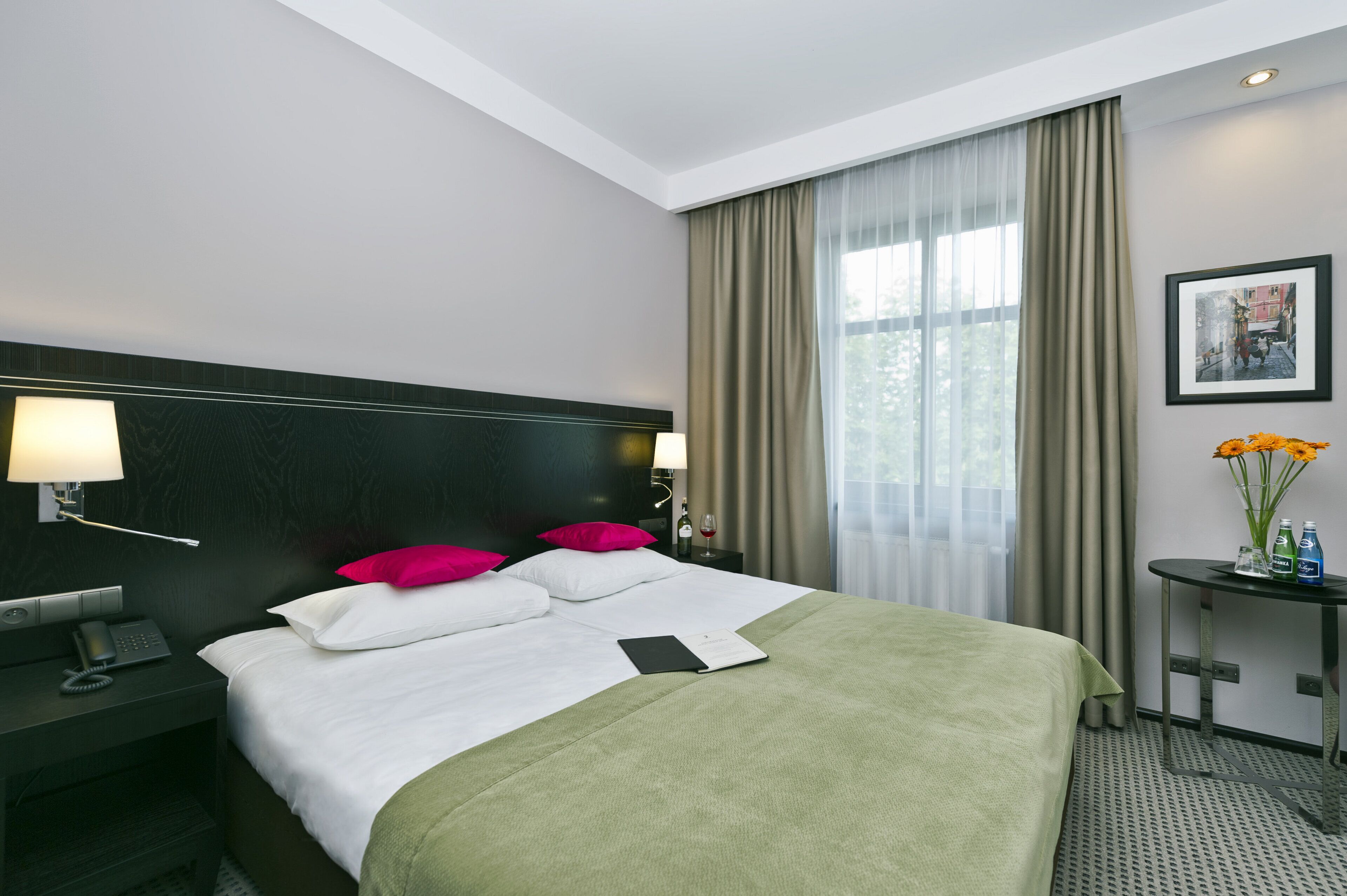 Un cuarto de hotel con una cama grande, una mesita de noche con una lámpara, una pequeña mesa con un florero y un cuadro en la pared.