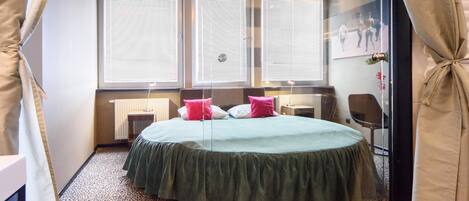 A bedroom with a round bed, a radiator, and three windows with blinds.
