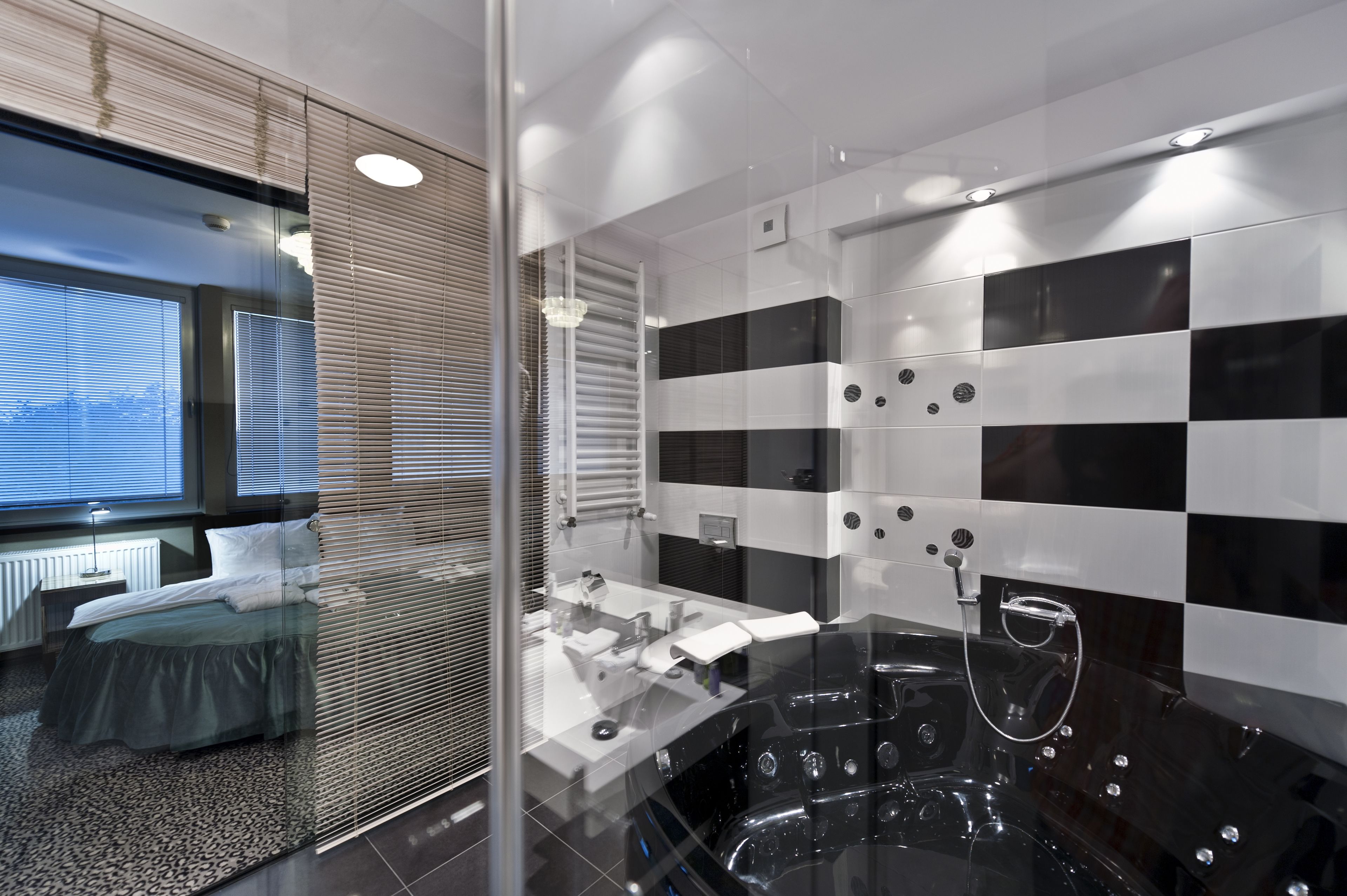 A modern bathroom with a glass partition, a black and white checkered wall, a large bathtub, and a visible window with blinds.