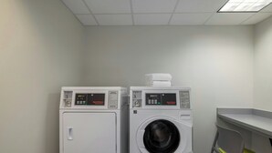 Een wasserette met twee witte industriële drogers en een wasmachine, een stoel en een kleine tafel.