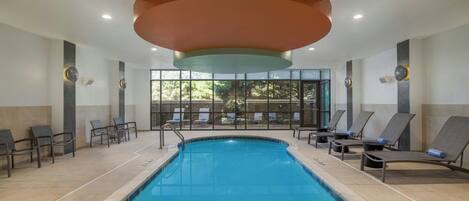 A swimming pool with lounge chairs and a view of trees through large windows.