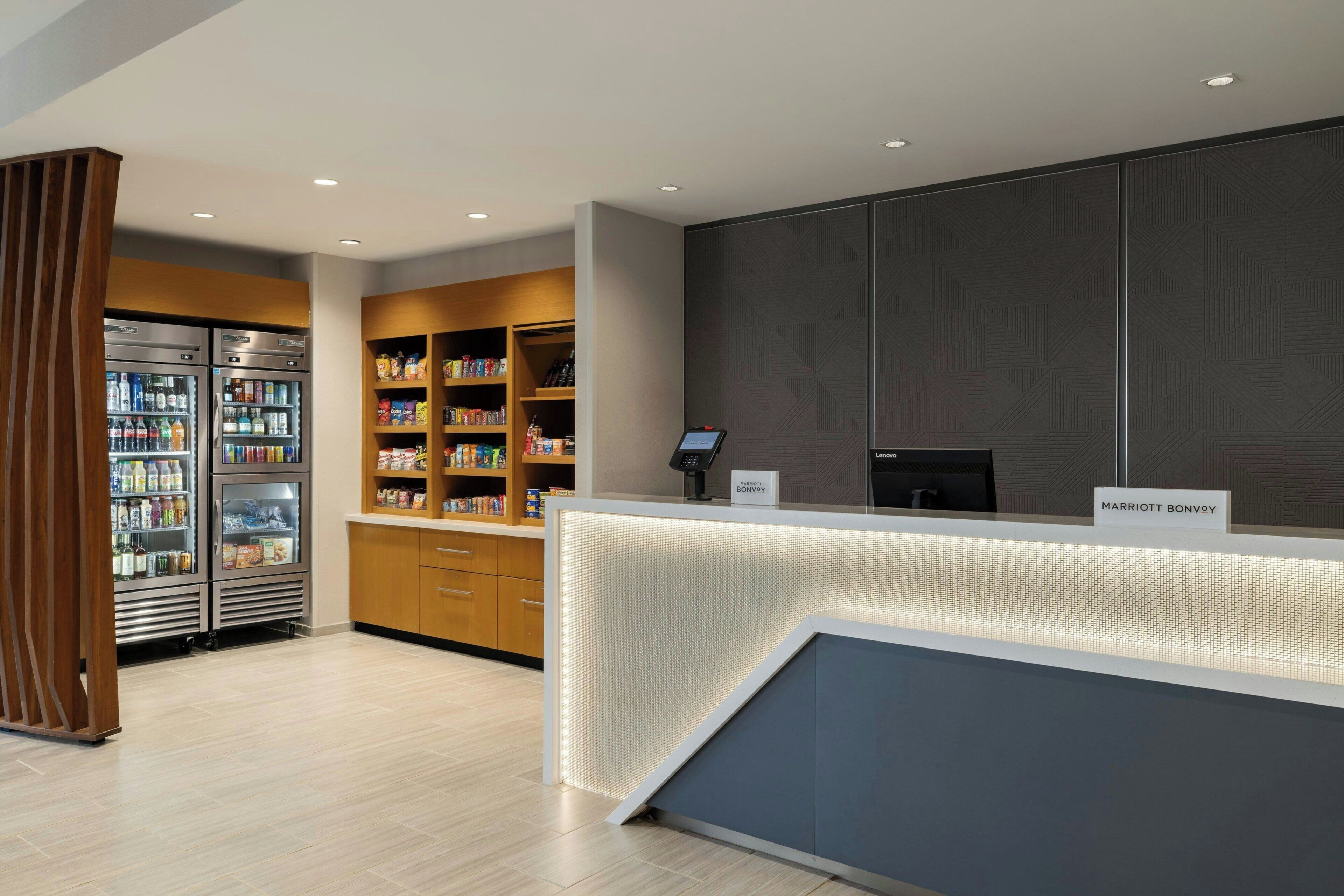 Un espace d’accueil d’hôtel au design moderne, comprenant un comptoir d’accueil, un kiosque en libre-service et un réfrigérateur rempli de boissons.